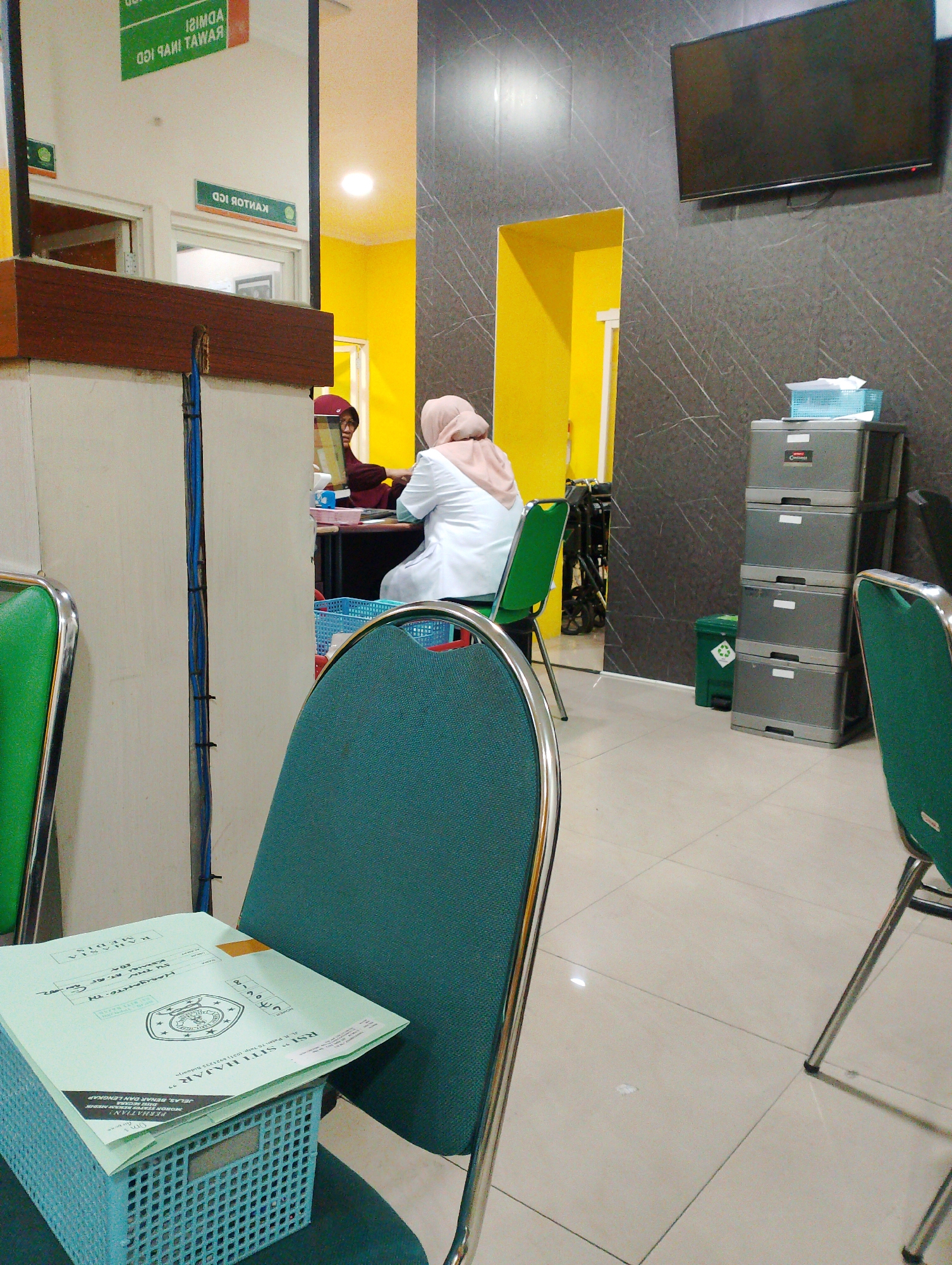 The image depicts an indoor scene at a medical facility in Sidoarjo, Indonesia. In the background, two medical personnel are visible behind a counter. One is wearing a white medical coat and a light pink hijab, while the other is wearing a dark red uniform and a red headscarf. They appear to be interacting with each other or with something on the counter. A television is mounted on the wall above a dark grey tiled surface. To the right, a stack of grey plastic drawers and a green recycling bin are present. In the foreground, a teal chair is prominent, with a light green folder containing medical documents resting on a blue plastic basket placed on another teal chair. The floor is tiled with light beige tiles. The overall environment suggests an administrative or waiting area within a hospital or clinic.