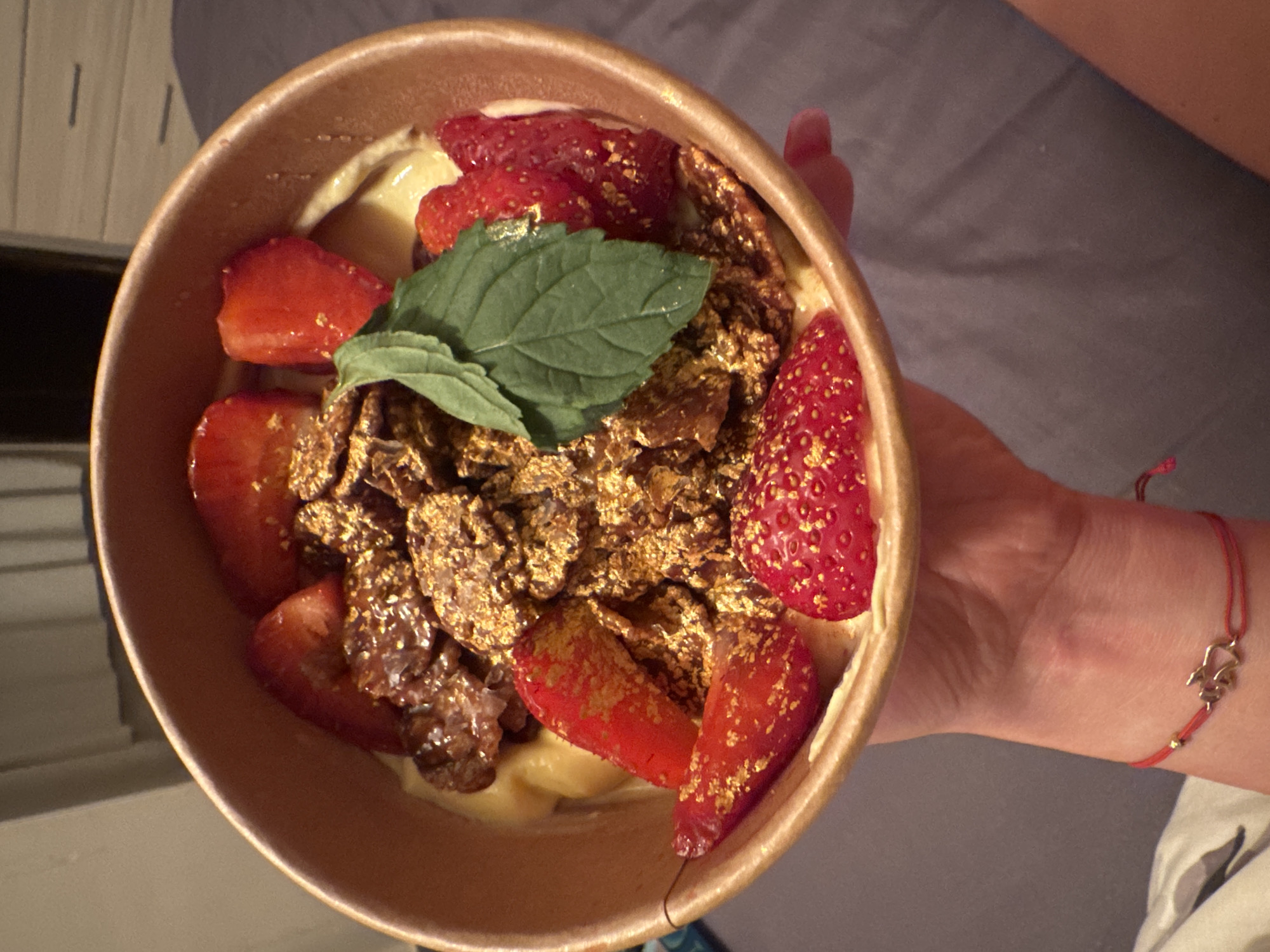 A top-down, close-up shot depicts a person holding a bowl of food, possibly in Varna, Bulgaria. The food appears to be a dessert consisting of sliced strawberries, granola (or a similar topping), a creamy substance (possibly yogurt or custard), and a couple of green leaves, likely mint. The strawberries and granola have a gold dusting. The bowl is held in the person's left hand, with part of their wrist visible, adorned with a red string bracelet featuring a gold charm. The background includes a glimpse of an interior space with white blinds and a gray surface.