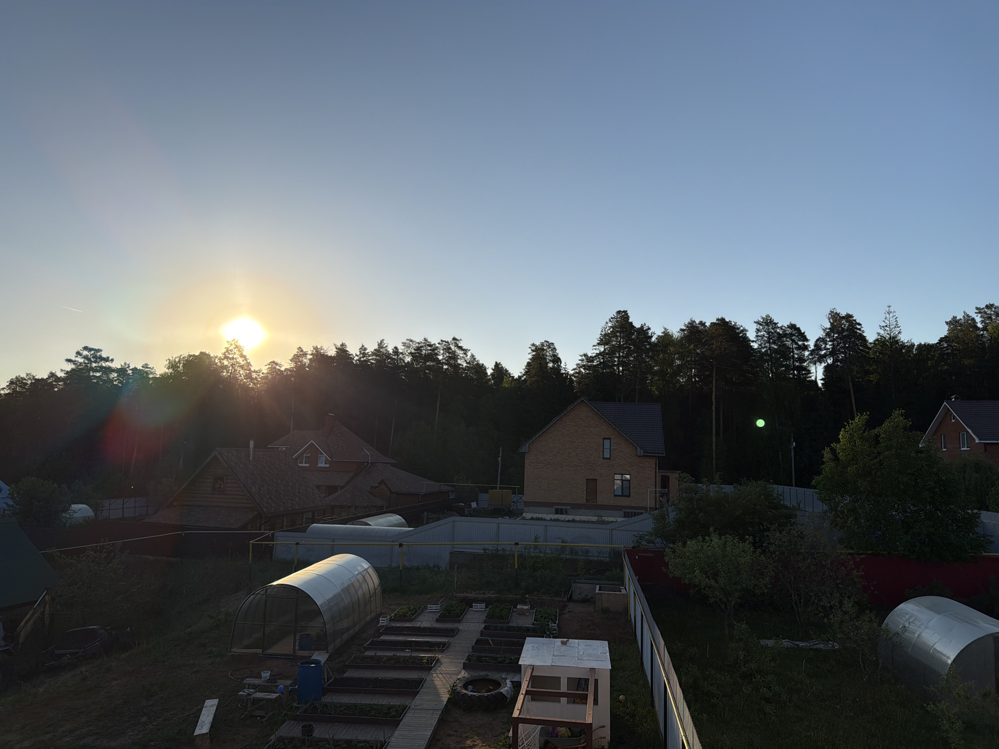 The image depicts a rural scene in the city of Chaikovsky, Russia. It is daytime, with the sun low on the horizon, partially obscured by trees. The sky is clear and blue. In the middle ground are several residential buildings constructed of wood and brick, with pitched roofs. A metal fence encloses the properties. A garden is in the foreground, with raised garden beds, two greenhouses, and a small, partially constructed structure. Dense trees are visible in the background. There are no people visible in the image.