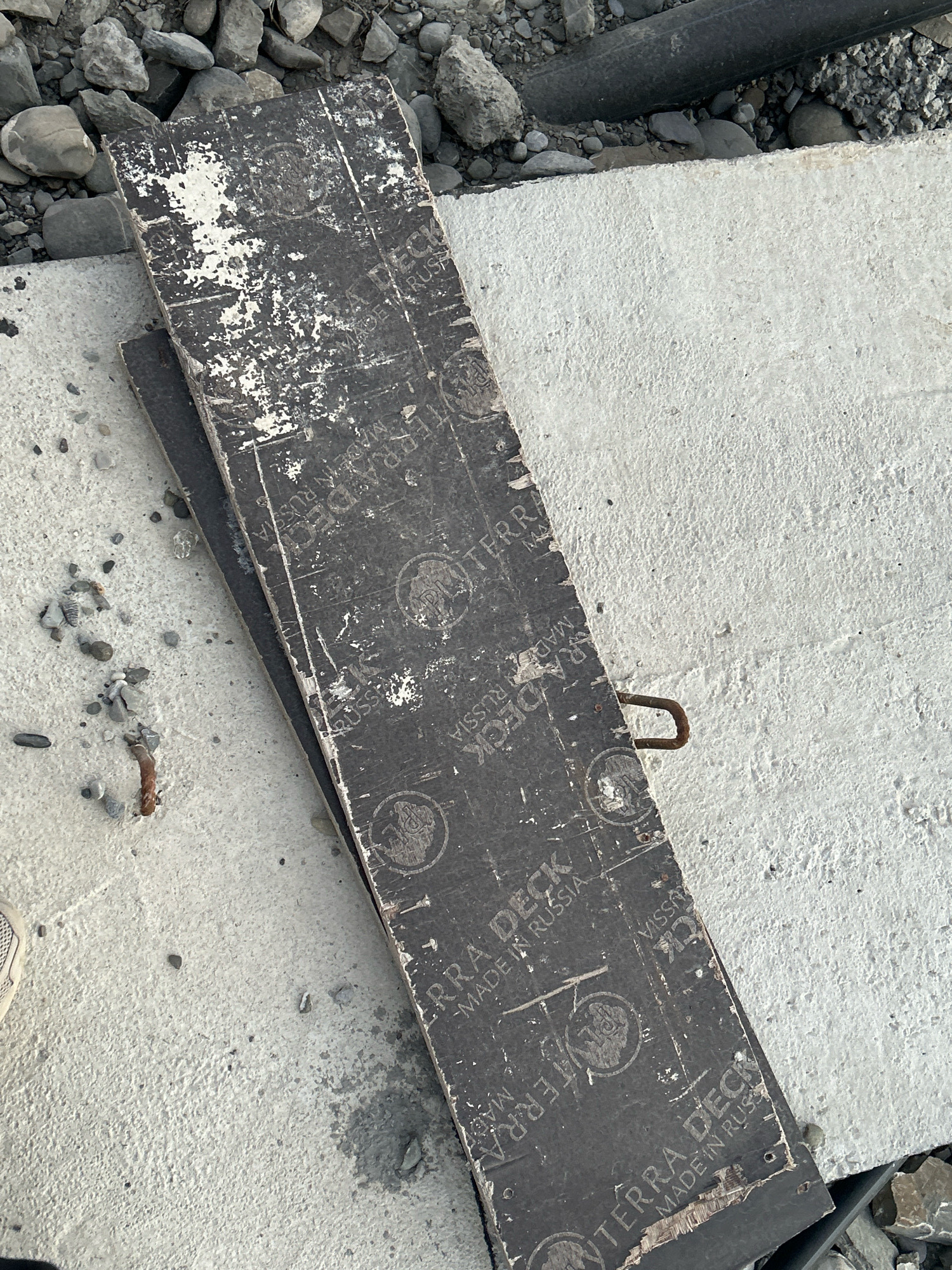 A close-up, overhead shot captures construction materials on an outdoor surface. Two dark, weathered wooden planks are layered diagonally on top of a rough, light-colored concrete slab. The top plank is distressed, with white paint peeling off and visible text that includes "TERRA DECK" and "MADE IN RUSSIA." Several circular stamps with an image inside are also visible on the planks. A rusty U-shaped metal hook protrudes from the edge of the top plank and rests on the concrete. The background consists of gray gravel and rocks, with a dark, cylindrical object, possibly a pipe, also present. A small section of a white shoe is visible in the lower left corner. The scene appears to be in an industrial or construction area, with no people actively present in the frame. The location is identified as Pavleuri, Georgia.