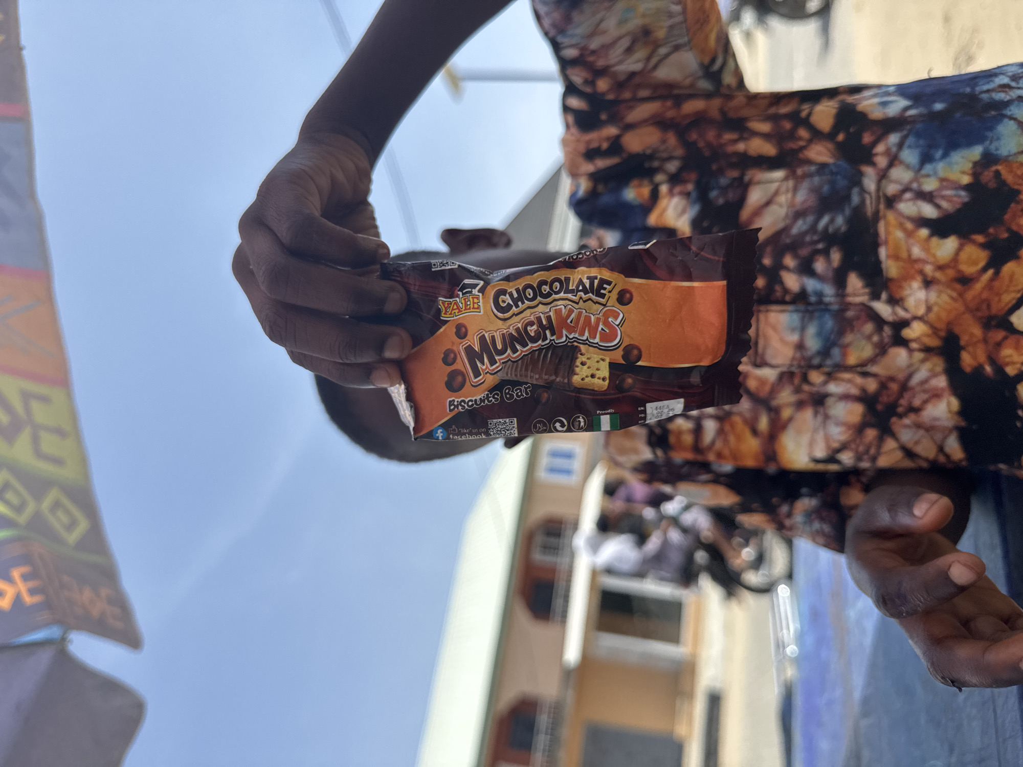 A person holds a "Yale Chocolate Munchkins Biscuits Bar" in their left hand. The packaging features chocolate-covered biscuits and the Nigerian flag, with manufacturing and expiration dates visible. The person is wearing a patterned shirt. The background shows a building with windows and a light blue sky, and what appears to be a bicycle. The location is Badagry, Nigeria, based on the product's flag, the buildings, and the clothing of the person.