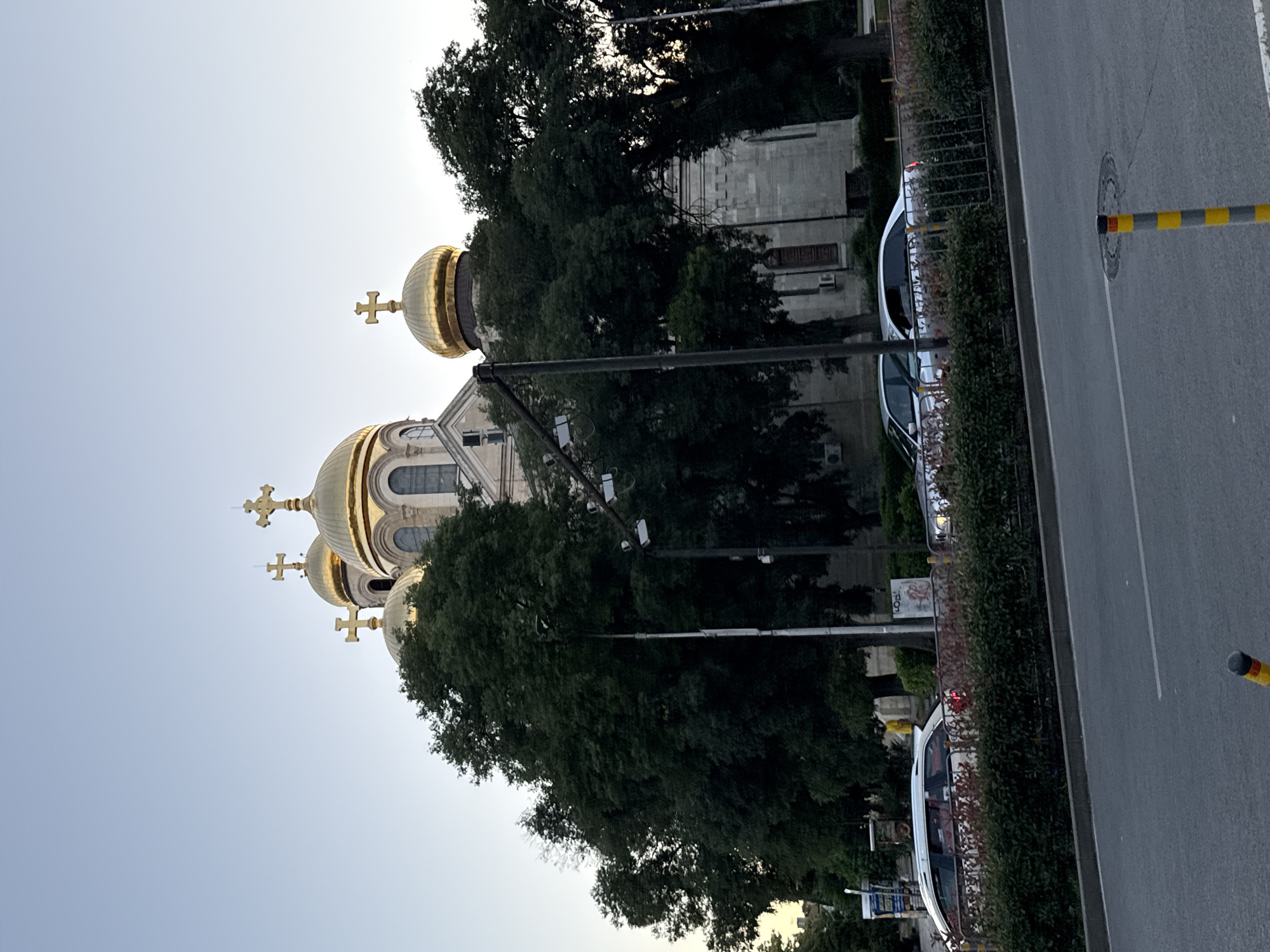 An exterior, eye-level, vertical, medium shot captures the golden domes of a cathedral peeking through dark green trees in an urban setting. The sky above is a pale, clear blue. In the foreground, a paved road with a yellow and black striped post is visible, separated by a low hedge and a metal fence from a sidewalk where several cars are parked or driving. The architecture of the cathedral is ornate, with arched windows and gold crosses atop its domes. The presence of traffic lights, streetlights, and air conditioning units on the building suggests a functional urban environment. The overall impression is a juxtaposition of sacred architecture and everyday city life.