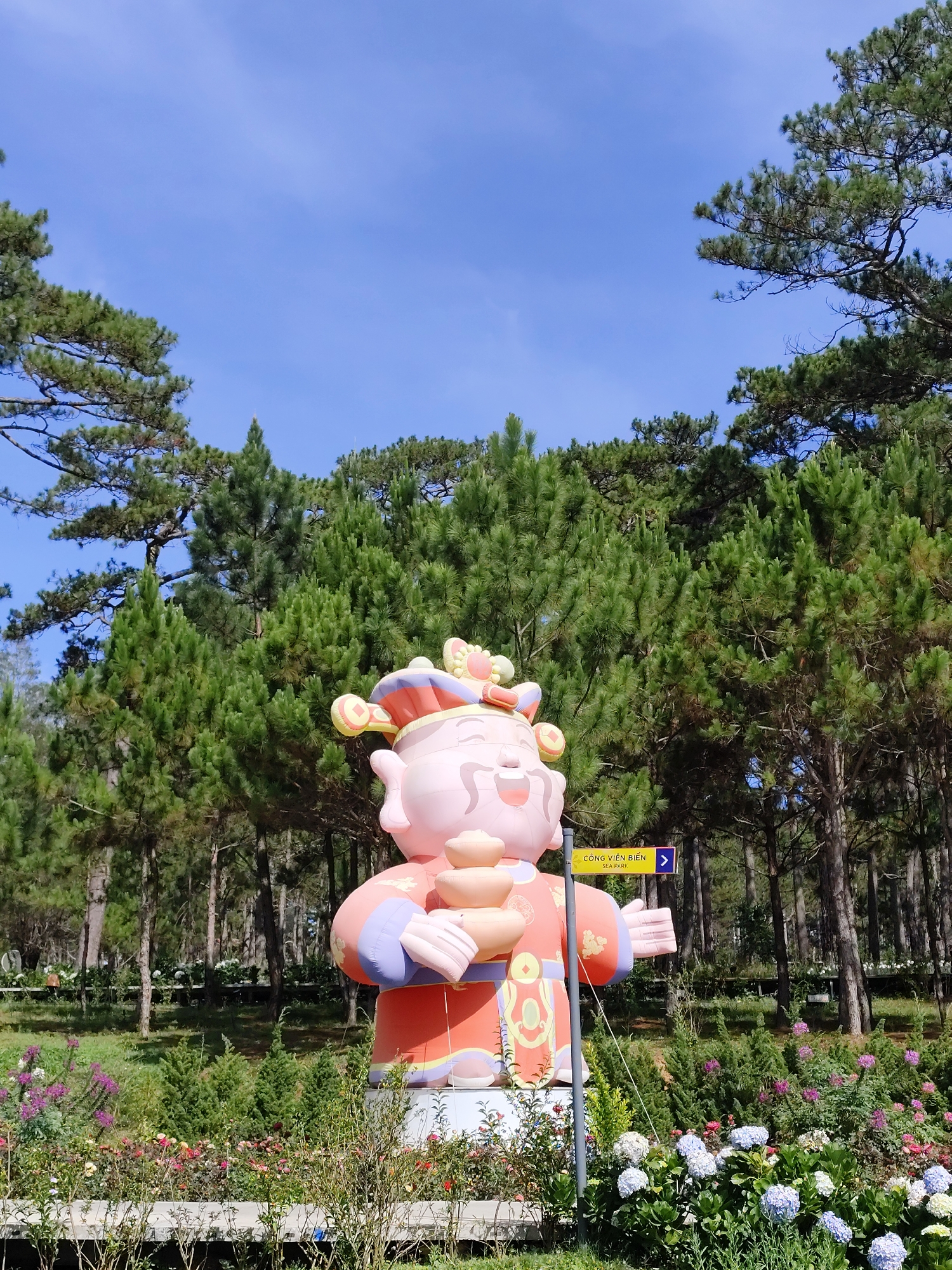 The image presents a vibrant, outdoor scene in Lâm Viên - Đà Lạt, Vietnam, featuring a large, colorful, inflatable sculpture of a traditionally dressed figure standing in a park setting. The figure, rendered in shades of pink, orange, and blue, is holding what appears to be golden ingots. A yellow sign with blue text and an arrow pointing right, marked "CÔNG VIÊN BIỂN SEA PARK," is positioned near the sculpture.
Behind the statue and sign, a dense forest of tall, green pine trees reaches toward a clear, blue sky. In the foreground, a flowerbed blooms with a variety of plants, including hydrangeas in shades of white and blue, interspersed with other colorful flowers. The scene is bathed in natural light, suggesting a clear, sunny day.