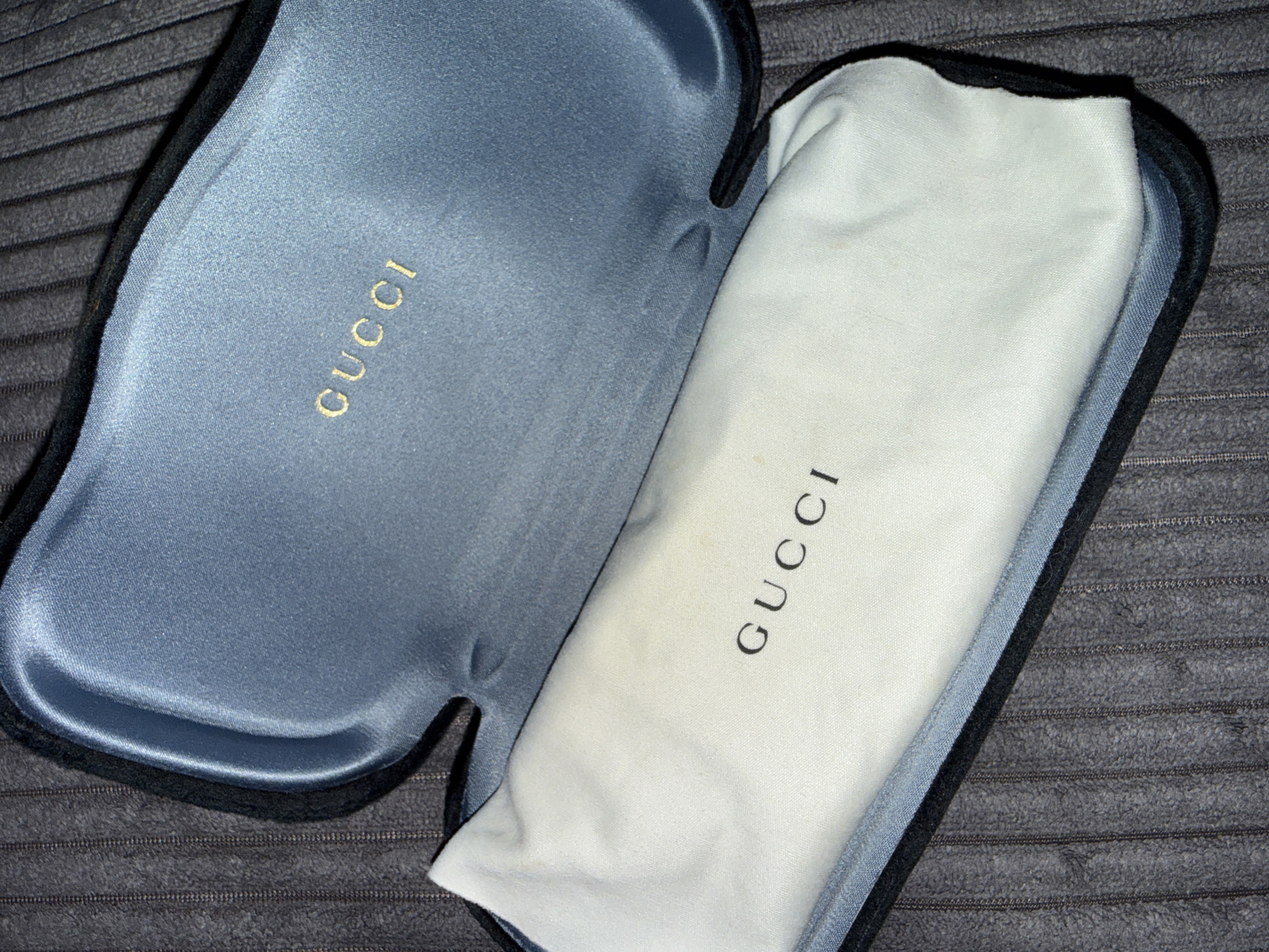 The image shows an open, dark blue, velvet-lined sunglasses case with a white fabric pouch inside. The Gucci logo is visible in gold lettering on the inside of the case and in black lettering on the white pouch. The case is resting on a dark gray, ribbed surface, possibly a textured fabric or carpet. There are no people or animals present in the image. The lighting appears to be from above, casting shadows within the case. The scene is a close-up, overhead shot. There is no information in the image to suggest a location in Varna, Bulgaria.