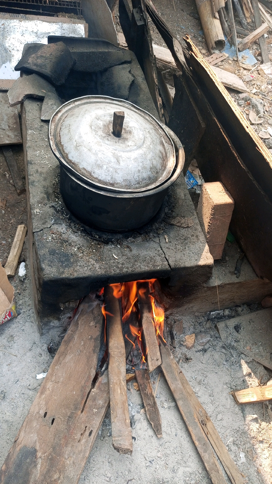 In Mangliawan, Indonesia, a traditional clay stove with a metal pot is seen in use. The stove is made of aged, cracked clay, with visible remnants of soot and ash. A fire burns beneath the pot, with bright orange flames licking the bottom. The pot is covered with a metal lid, topped with a small wooden handle. Scattered around the stove are pieces of wood, some of which are fueling the fire, and other debris, suggesting an outdoor or rustic setting. The background shows more discarded materials, including pieces of wood and what appears to be urban detritus, indicating a possibly impoverished or informal living environment.