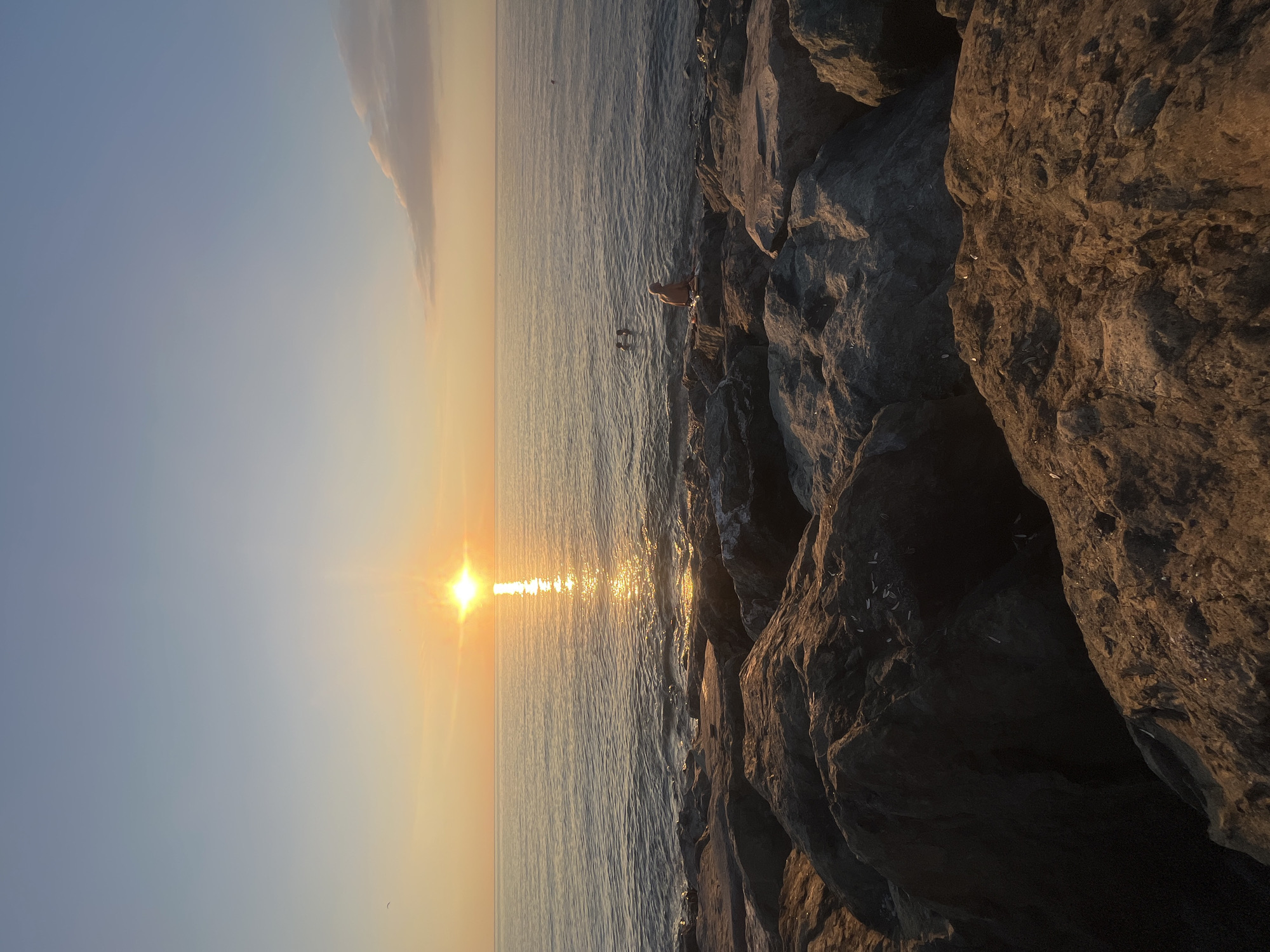 The image captures a serene sunset scene on a rocky coastline, likely in Batumi, Georgia. The sun, a radiant orb with a distinct sunburst effect, is low on the horizon, casting a golden glow across the tranquil sea. The water reflects the warm hues of the sunset, creating a shimmering path of light that stretches towards the shore.
The foreground is dominated by large, textured rocks, typical of a coastal environment. These rocks, dark and rugged, provide a stark contrast to the soft, ethereal colors of the sky and sea. A few small white specks, possibly feathers or debris, are scattered across the rock surfaces.
In the water, near the shore, two figures are visible. One person appears to be standing in the shallow water, while another is partially submerged. Their presence adds a sense of life and activity to the otherwise quiet landscape. The overall mood is peaceful and contemplative, evoking a feeling of calm and natural beauty. The time of day is clearly sunset, with the warm, low-angled light characteristic of this period. The weather appears calm, with still water and a clear sky, save for a wispy cloud formation in the distance.