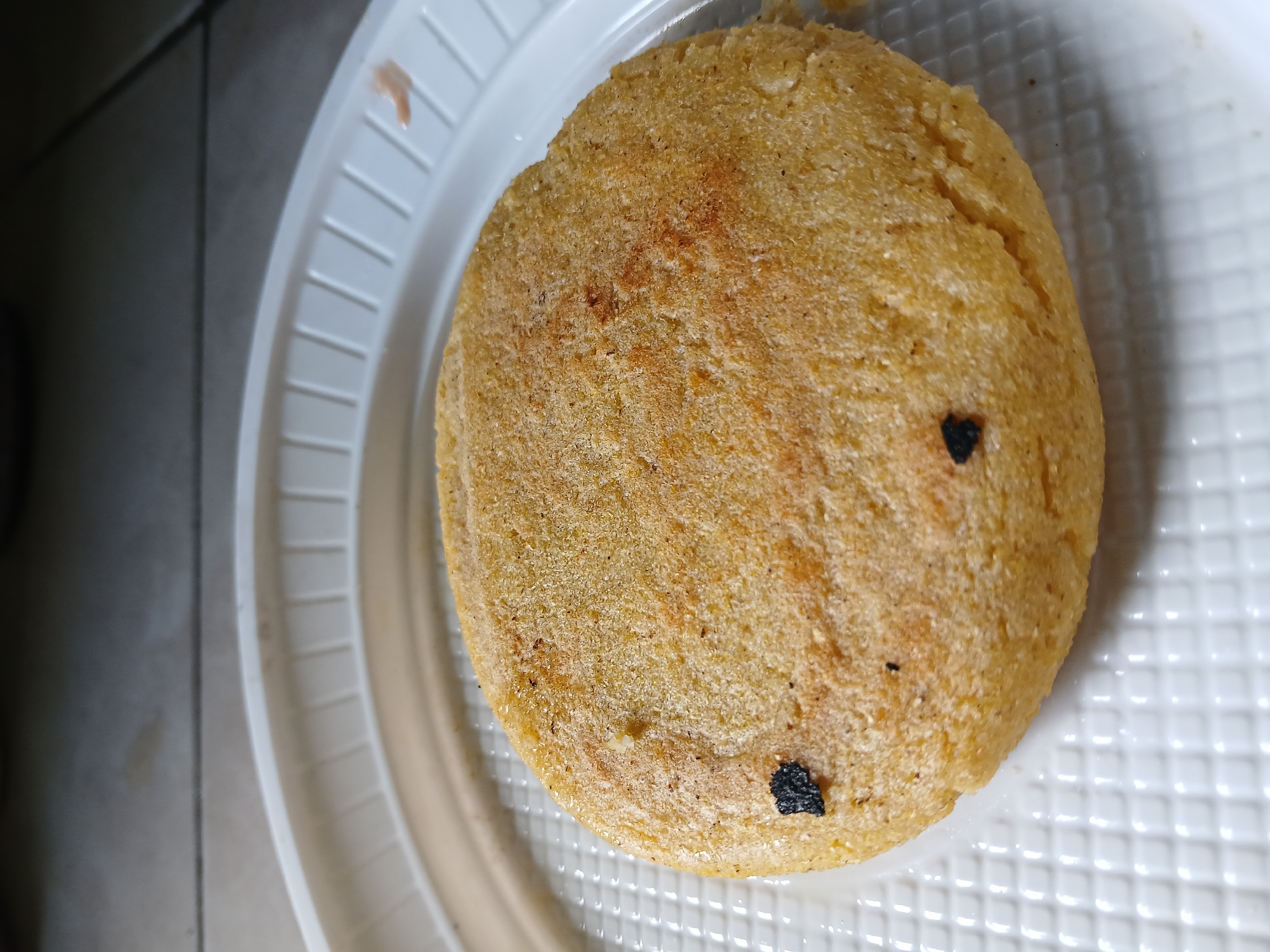 The image shows a single arepa, a type of flatbread made from ground maize dough, placed on a white plastic plate. The arepa is golden brown in color, with some darker spots that indicate grilling or toasting. There are also two black specks adhered to the flatbread. The plate is a disposable type, with a ridged pattern. The background is blurry and shows part of what appears to be a countertop. The image provides no direct evidence of a location in Juan Griego, Venezuela.
