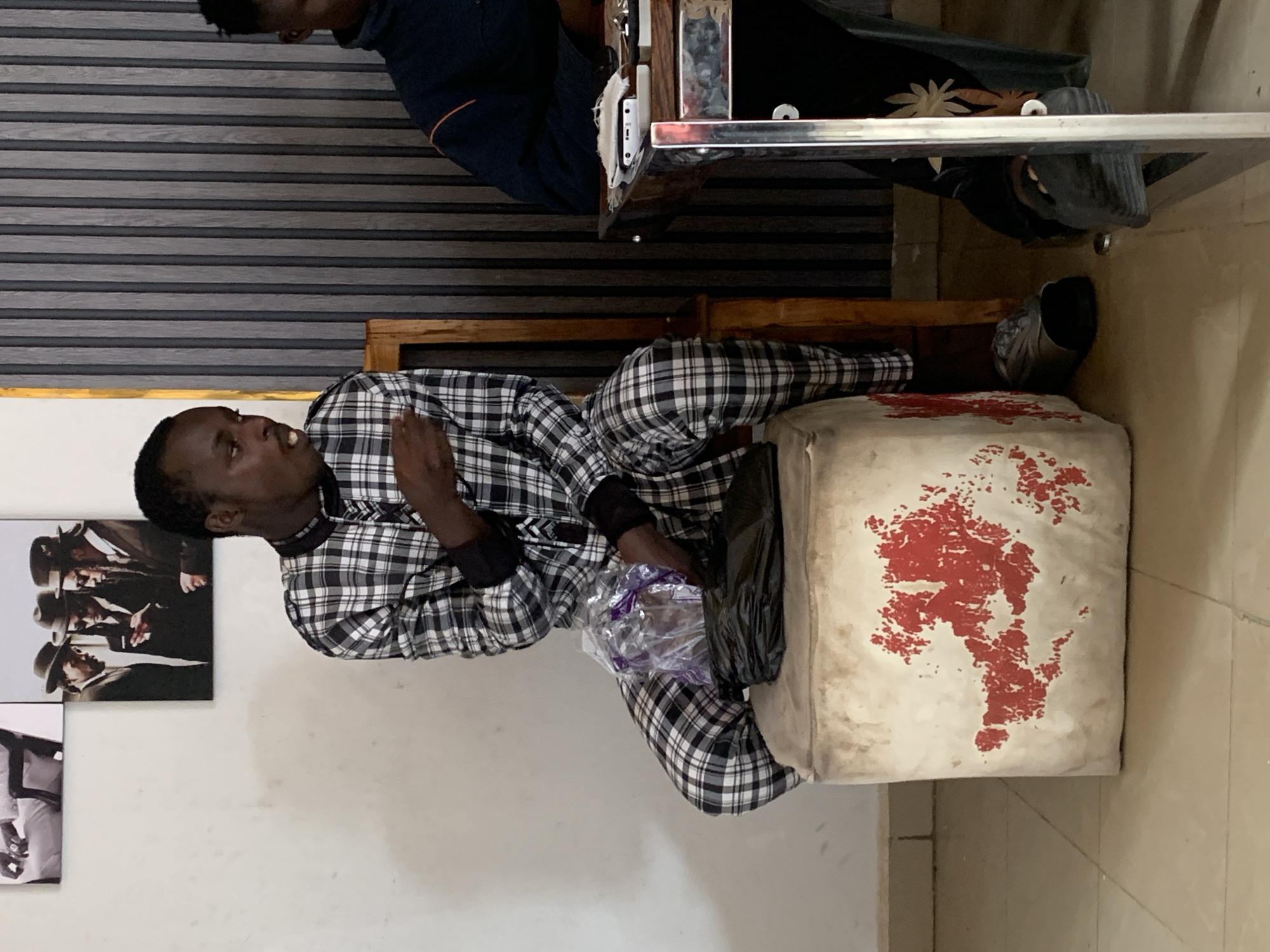 A man wearing a plaid pajama set is sitting on a worn, square ottoman with red stains. He is holding a clear plastic bag and a black bag. He appears to be looking up and to the left, gesturing with his right hand. To the left of the man are framed black and white pictures on a white wall. In the background, there are horizontal striped blinds. Another person is partially visible at the top of the image, sitting at a table. The image appears to be taken indoors. The specific location within the city of Jos, Nigeria is indeterminable from the image.
