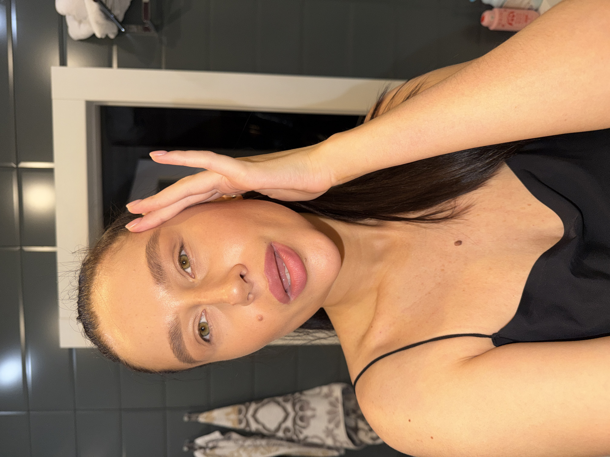 The image, originally rotated 90 degrees clockwise, captures a close-up selfie of a young woman. She is looking directly at the camera with a calm and slightly posed expression. Her hair is dark and appears to be pulled back, framing her face. She has prominent, well-groomed eyebrows and light-colored eyes, appearing green or hazel. Her lips are slightly parted, showing a natural pink shade, and her skin is fair with visible freckles and several moles on her face and body, including one prominent mole below her right eye and another on her upper chest. Her right hand is raised to her forehead, with her fingers gently resting on her temple, and her nails are manicured with a light, natural-toned polish. She is wearing a black top or dress with a thin spaghetti strap visible over her left shoulder.
The background suggests an indoor setting, possibly a bathroom or a similar tiled space. Dark grey or black square tiles cover a wall behind her. A white architectural element, possibly a mirror or door frame, is prominent behind her left side. In the upper right corner, a pink bottle, consistent with a personal care product like shampoo or lotion, is visible, along with some white fabric, possibly a towel. A patterned towel or fabric is also visible in the lower background. The lighting is even, illuminating her face and upper body. No visible text is clearly discernible on the subject or in the immediate background objects. The photo's context is Khimki, Russia.