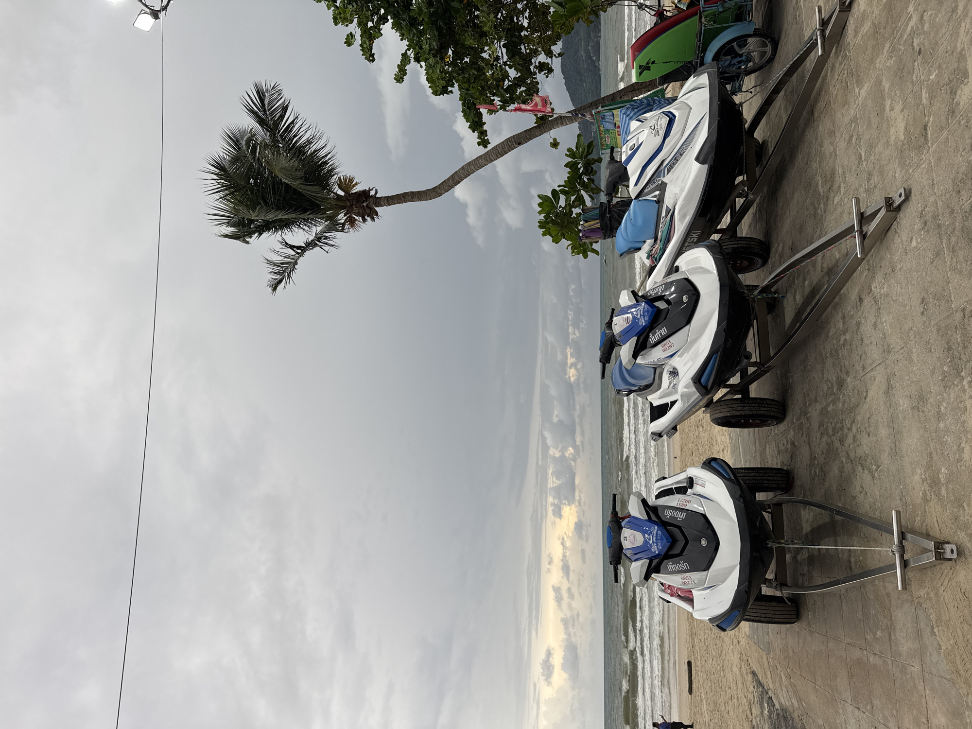 In Patong, Thailand, three jet skis are parked on a paved area next to a sandy beach. The jet skis are white and blue, with one jet ski to the left displaying the number "3" on its front. They are all on trailers with wheels, ready for use on the water. The ocean is visible in the background with moderate waves. A single person, appearing to be a man in a dark shirt and pants, stands on the beach to the left of the jet skis. A palm tree leans over the scene, and lush green foliage is present on the right. The sky is overcast with a light gray hue.