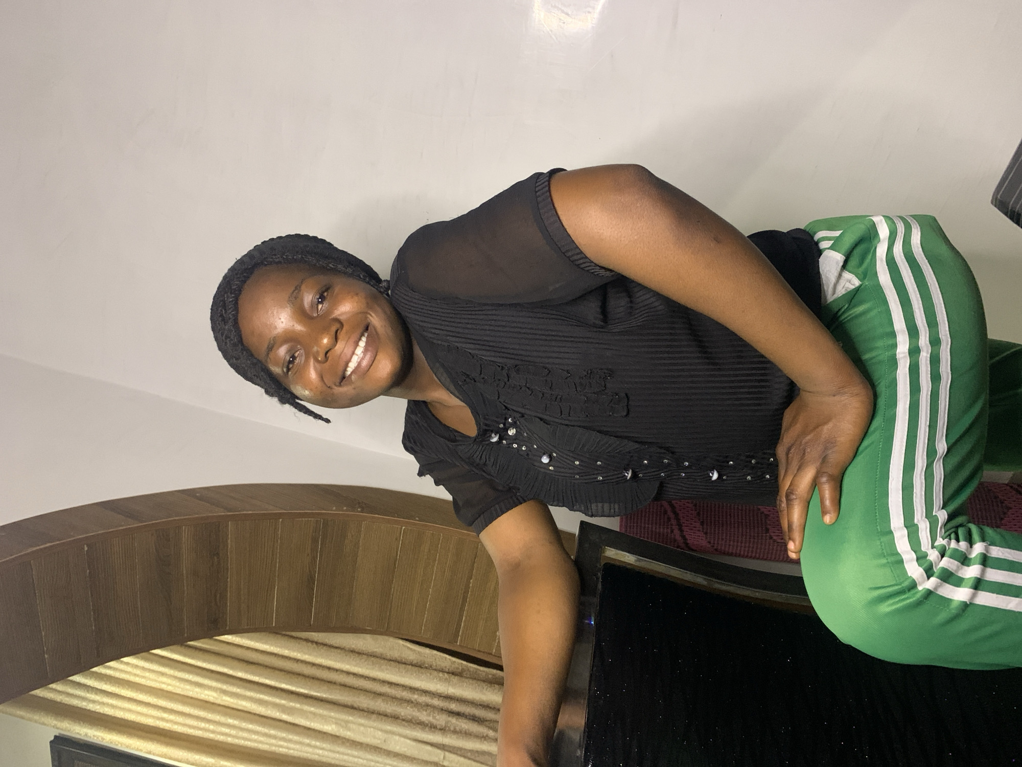 A young Black woman with dark skin, dark braided hair, and a bright smile is captured in a medium shot. She is wearing a black, sheer, short-sleeved blouse with ruffled detailing down the front and adorned with small, sparkly embellishments. Her lower body is clad in vibrant green track pants with three white stripes along the sides. She is seated, leaning forward with her right arm resting on her knee. The background features a plain white wall and a curved wooden structure with vertical slats, partially obscuring a golden curtain. To the left of the woman, a dark, reflective surface, possibly a table or desk, is visible. The lighting is soft, creating subtle shadows on the wall and the woman's face. The overall impression is that of a casual portrait, possibly taken indoors in a residential setting in Jos, Nigeria.