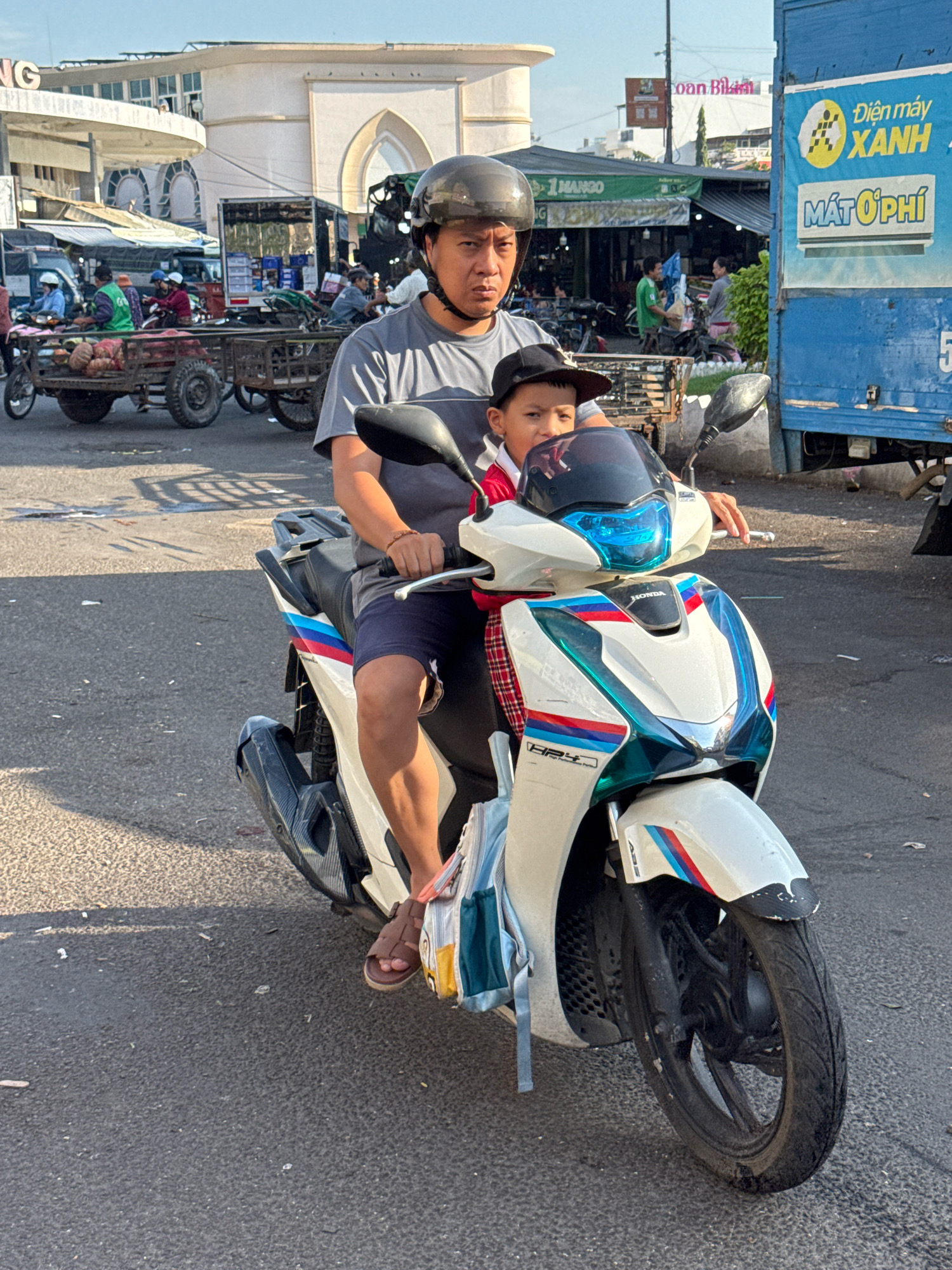 In Nha Trang, Vietnam, a man and a young boy ride a white Honda scooter on an asphalt road. The man, wearing a gray t-shirt and a black helmet, is driving. The boy, wearing a black baseball cap and a red and white plaid shirt, sits in front of him. The scooter has blue, red, and white racing stripes and a black exhaust pipe. In the background, a blue truck with Vietnamese writing is visible, along with market stalls and other vehicles. The sky is clear and bright, suggesting it is daytime.