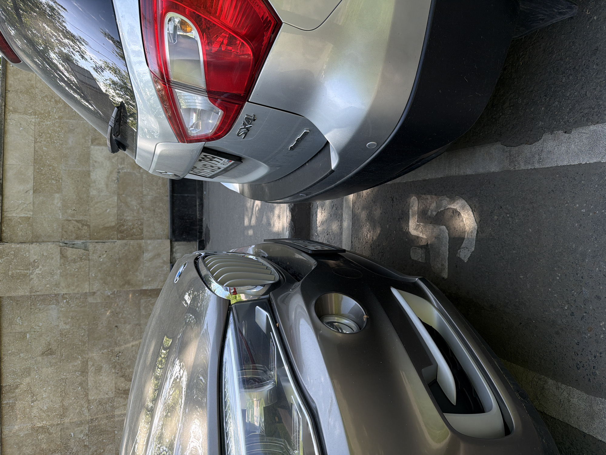 The image shows the rear of a silver Suzuki SX4 and the front of a gray BMW parked next to each other in a parking spot. The parking spot is marked with the number "5" painted on the asphalt. The location is likely Moscow, Russia, based on the license plate seen on the Suzuki. The cars are parked in an outdoor environment with some trees visible.