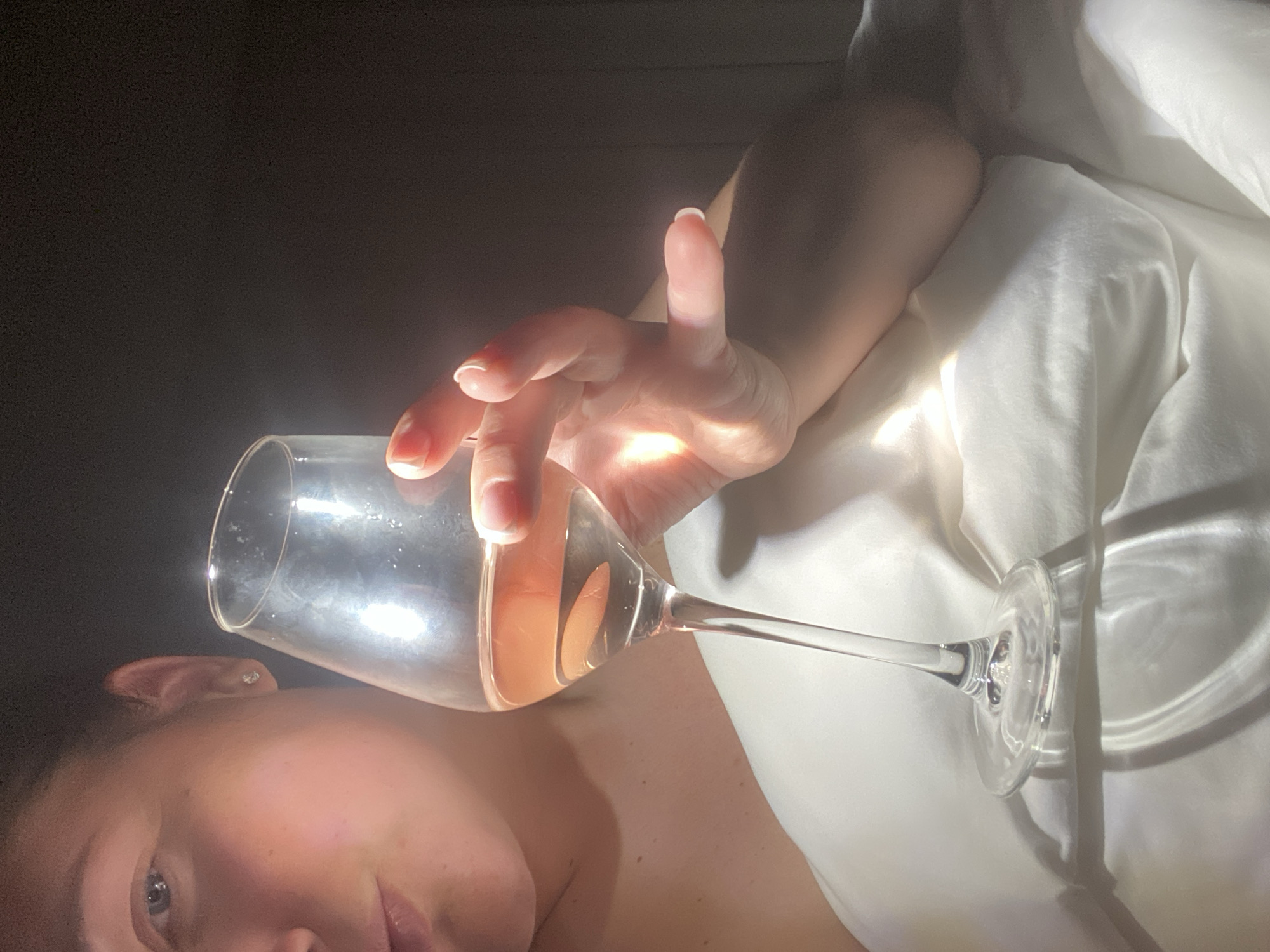 A wine glass partially filled with a pink liquid is held by a hand in a dimly lit room, illuminated by a bright light source casting stark shadows. The background consists of dark areas and what appears to be white fabric draped around, possibly bedsheets. A person's face, including the eye, cheek, and lips, is visible in the foreground. The photograph does not indicate a location in Sochi, Russia.