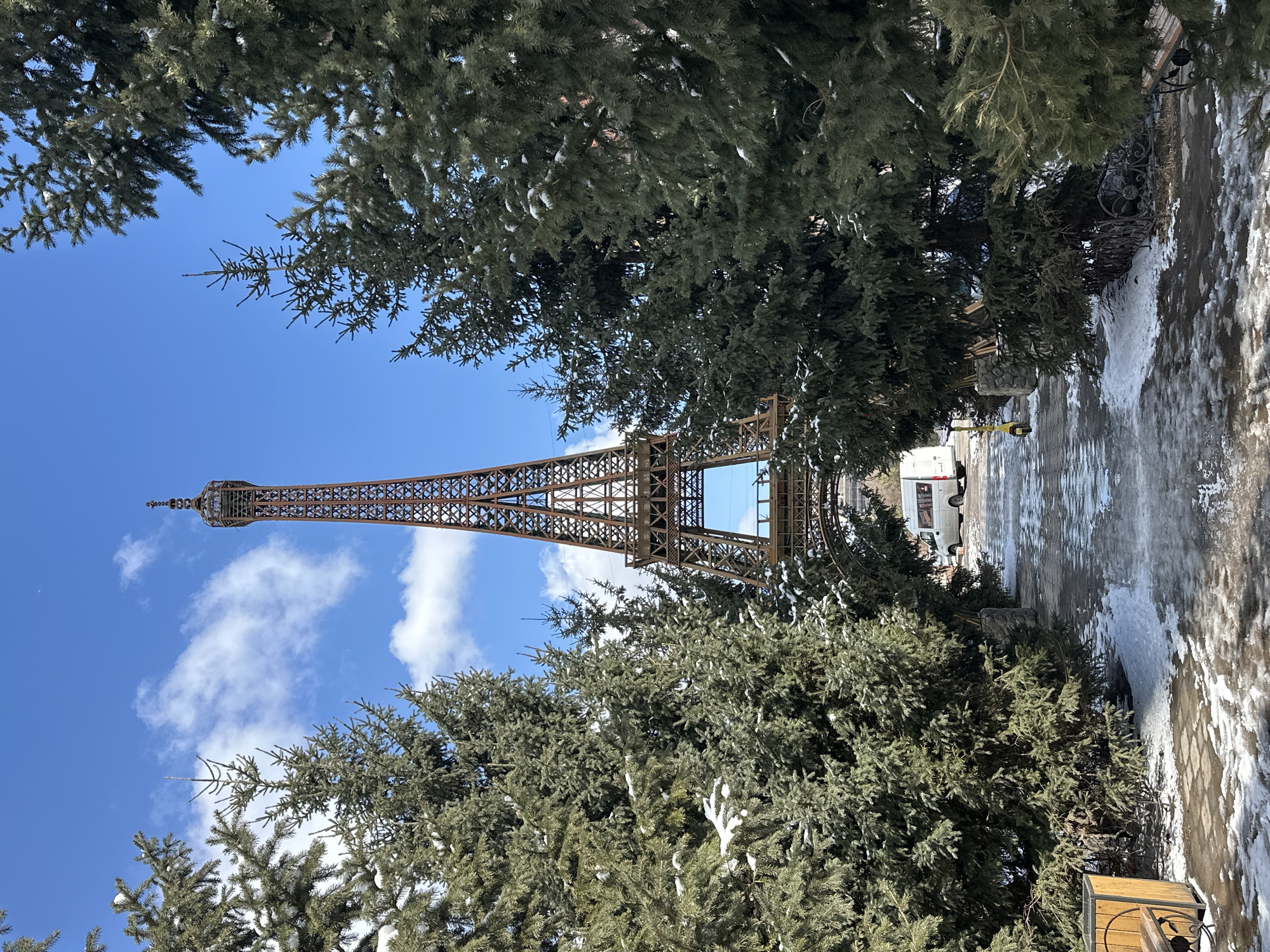 The image shows a view of a replica of the Eiffel Tower in Krasnoyarsk, Russia, framed by evergreen trees. The tower is constructed of brown metal lattice, and it extends upwards towards a blue sky dotted with white clouds. Some of the trees are dusted with snow. A paved path runs alongside the trees and the tower, with patches of snow and ice. A white van is parked in the background.