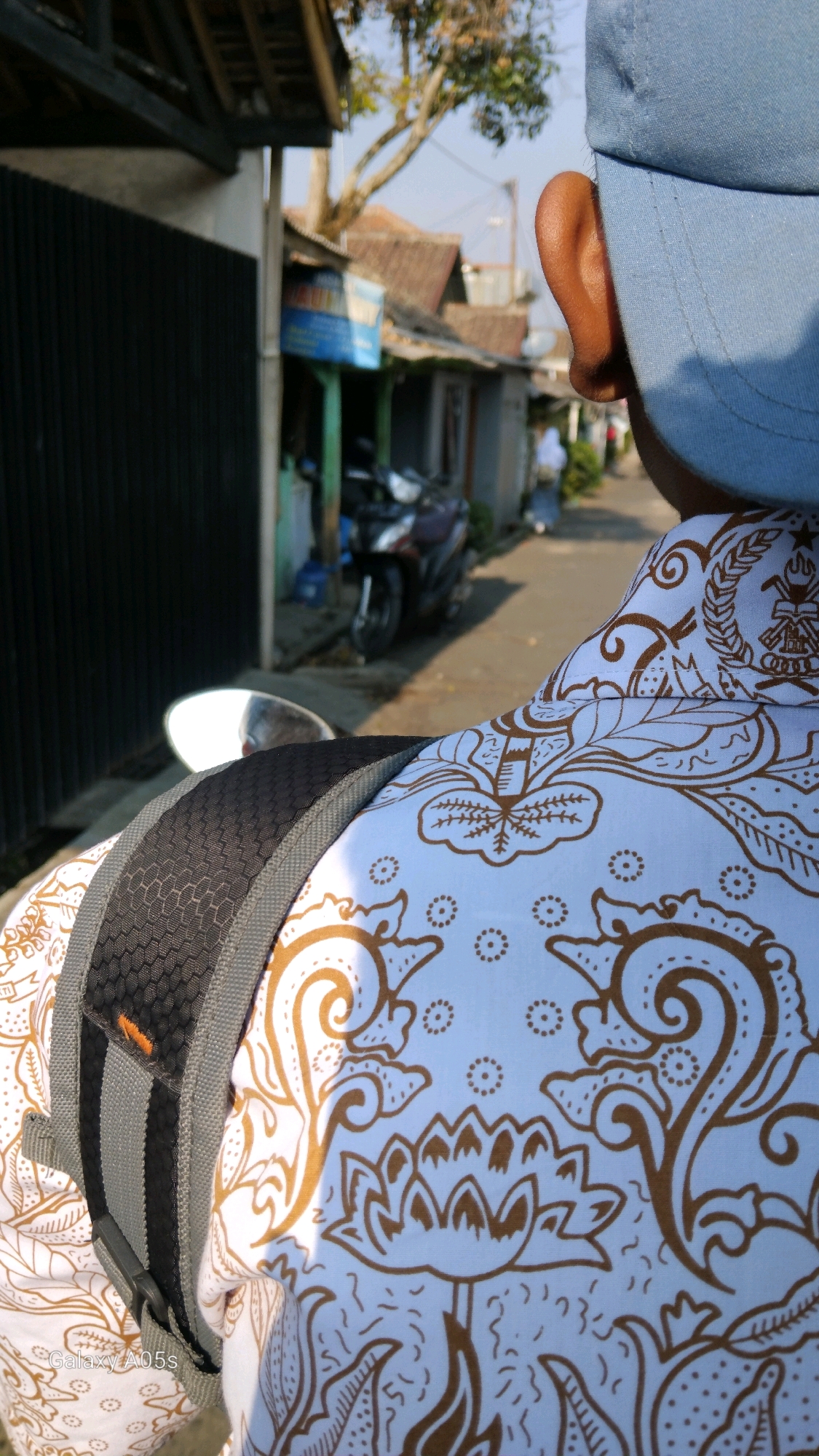 This is a close-up, over-the-shoulder shot of a person wearing a light blue baseball cap and a white shirt with a brown batik pattern. The person is viewed from behind and to the side, with their right ear visible. The batik pattern features stylized floral motifs and circular dots. A black backpack strap with gray accents is visible across the person's shoulder. In the background, a narrow street is lined with buildings, some with tin roofs. A motorcycle is parked on the side of the street. Sunlight casts shadows on the scene. The image appears to have been taken in an urban or semi-urban environment in Sumedang, Indonesia, likely during daylight hours. A watermark indicating "Galaxy A05s" is present in the lower left corner.