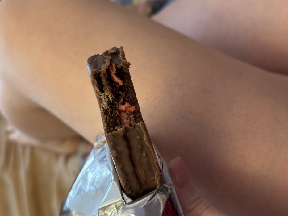A close-up, high-angle view depicts a partially consumed chocolate bar with a bite taken from its right end, revealing layered internal components. The bar is coated in dark brown chocolate, with an internal structure consisting of an upper dark brown layer, a vibrant orange-red filling, and a bottom dark brown layer. The item is held by a left hand, visible on the lower left, which has manicured nails with a light, opaque polish. The chocolate bar is partially encased in crinkled metallic silver foil packaging, visible on the left side of the frame, with a small red graphic detail near the bottom left edge of the packaging. The background consists primarily of human skin, specifically the upper thighs or legs of an individual, presenting a light-toned complexion with visible hair follicles. The lighting is soft and consistent, suggesting an indoor, casual setting. No discernible elements in the image provide information regarding a specific location within the city of Perm, Russia, or any other geographical location.