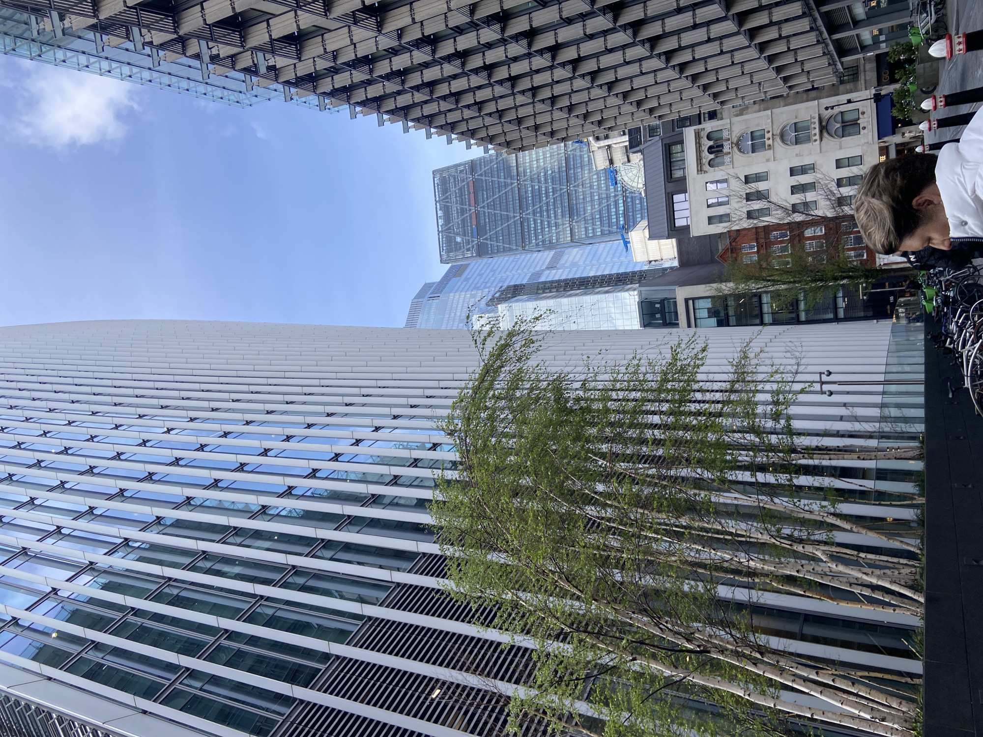 The image displays an upward-looking perspective of a dense urban environment, rotated 90 degrees clockwise. On the left, a tall, modern building features a facade of alternating horizontal white bands and reflective glass panels, with some sections of dark vertical slats. In front of this building, several slender trees with light green foliage grow upwards (to the right in the rotated view).
In the mid-ground, a cluster of modern skyscrapers with glass and metallic exteriors is visible, including one prominent building with a large, distinctive grid-like framework on its upper floors. To the right of these contemporary structures, a traditional, light-colored stone building with ornate arched windows and classic architectural details stands out, partially obscuring a smaller brick building below it. The sky, appearing on the top side of the rotated image, is a bright, clear blue with a few scattered white clouds.
On the far right edge of the frame, the head and shoulders of a person with light-colored hair, wearing a white jacket, are visible, looking downwards. Next to them, multiple bicycles are parked in public racks. Overhead (on the bottom side of the rotated image), a building's underside or upper structure overhangs the scene, characterized by a complex pattern of rectangular, possibly wooden or metallic, panels.
The scene depicts a sunny daytime moment. Visible text includes "GO..." and "COFFEE" on a dark building behind the traditional stone building, and "155" on a building situated below the traditional stone building. The image captures a blend of modern and traditional architecture in a bustling city setting.