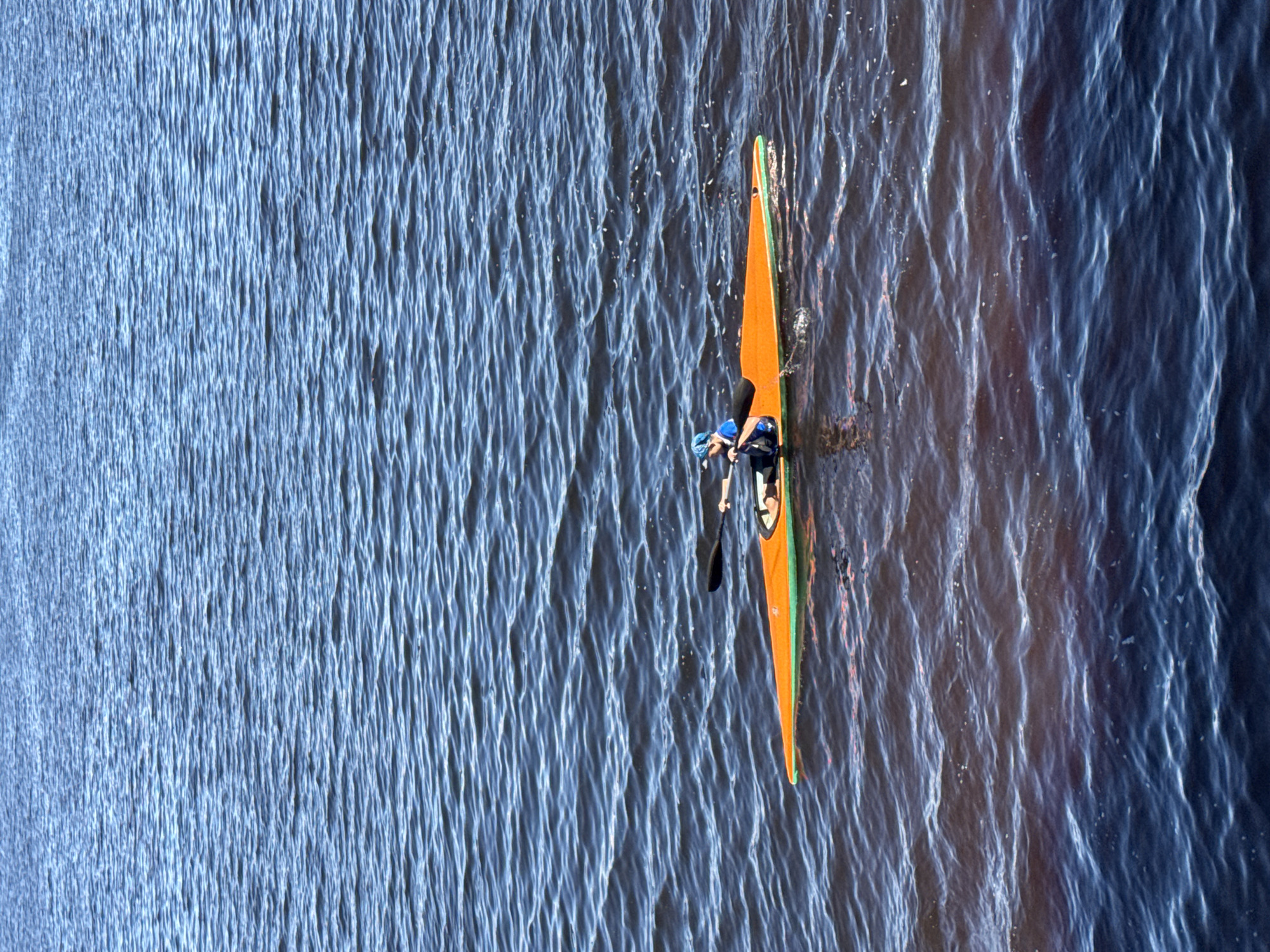 A person is kayaking on a body of water. The kayak is orange with a green trim. The person is wearing a blue shirt and a blue cap. The water is a deep blue with ripples and small white caps. The sky is not visible. The image appears to be taken from a slightly elevated position, looking down at the kayak. This scene is likely located in the city of Chaikovsky, Russia, known for its scenic water bodies and recreational activities, possibly along the Kama River or one of its tributaries.