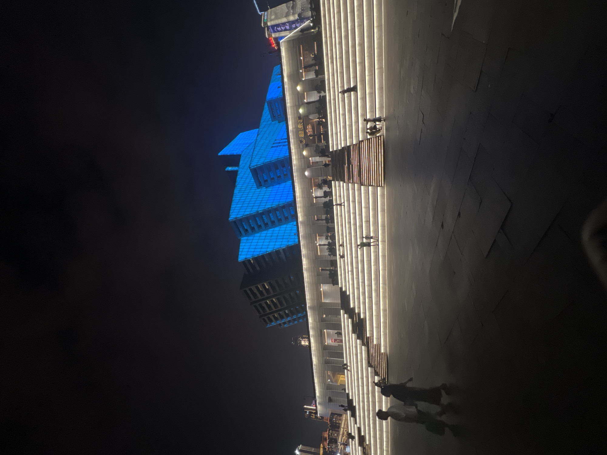 At night, a modern building with a glass facade illuminated in bright blue stands tall against a dark sky. In front of the building is a wide, multi-tiered staircase with illuminated steps, creating a luminous pathway. Several people are visible on the steps, some walking, others standing or sitting. Two individuals are walking in the foreground on a paved area. The architectural style of the building suggests it could be a cultural or entertainment venue. The location is identified as Jiangbeicheng Street, China.
