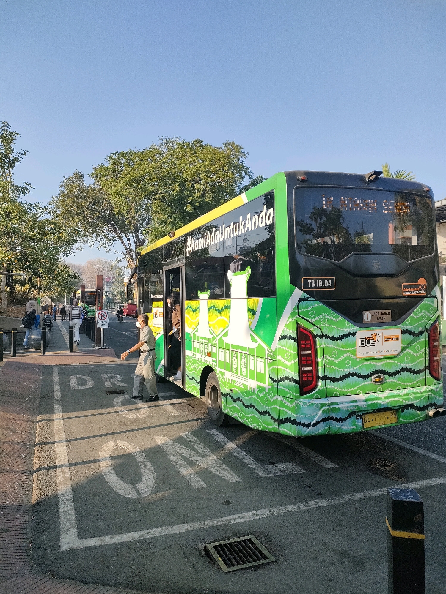 A bus with a green and white wave pattern is stopped at a bus stop. The bus is oriented towards the right side of the frame. The bus's rear is visible, displaying text in Indonesian that reads "#KamiAdaUntukAnda" and "TB 1B.04". Two young women are exiting the bus through the open door, and a male bus attendant in a light-colored uniform is standing by the door, facing them. A "No Parking" sign is visible on the left side of the road, near the sidewalk where several pedestrians are waiting. The road surface has painted white lines and markings, including the word "DRIVE" and "ONE" painted in large white letters, indicating a specific lane or zone. The environment appears to be an urban setting with trees lining the street and a clear blue sky overhead. A yellow license plate with text, likely the bus's registration, is visible on the rear of the bus. The scene suggests normal daily activity at a public transportation stop. The specific location within Banjarmasin, Indonesia, is not explicitly identifiable from the visual elements.