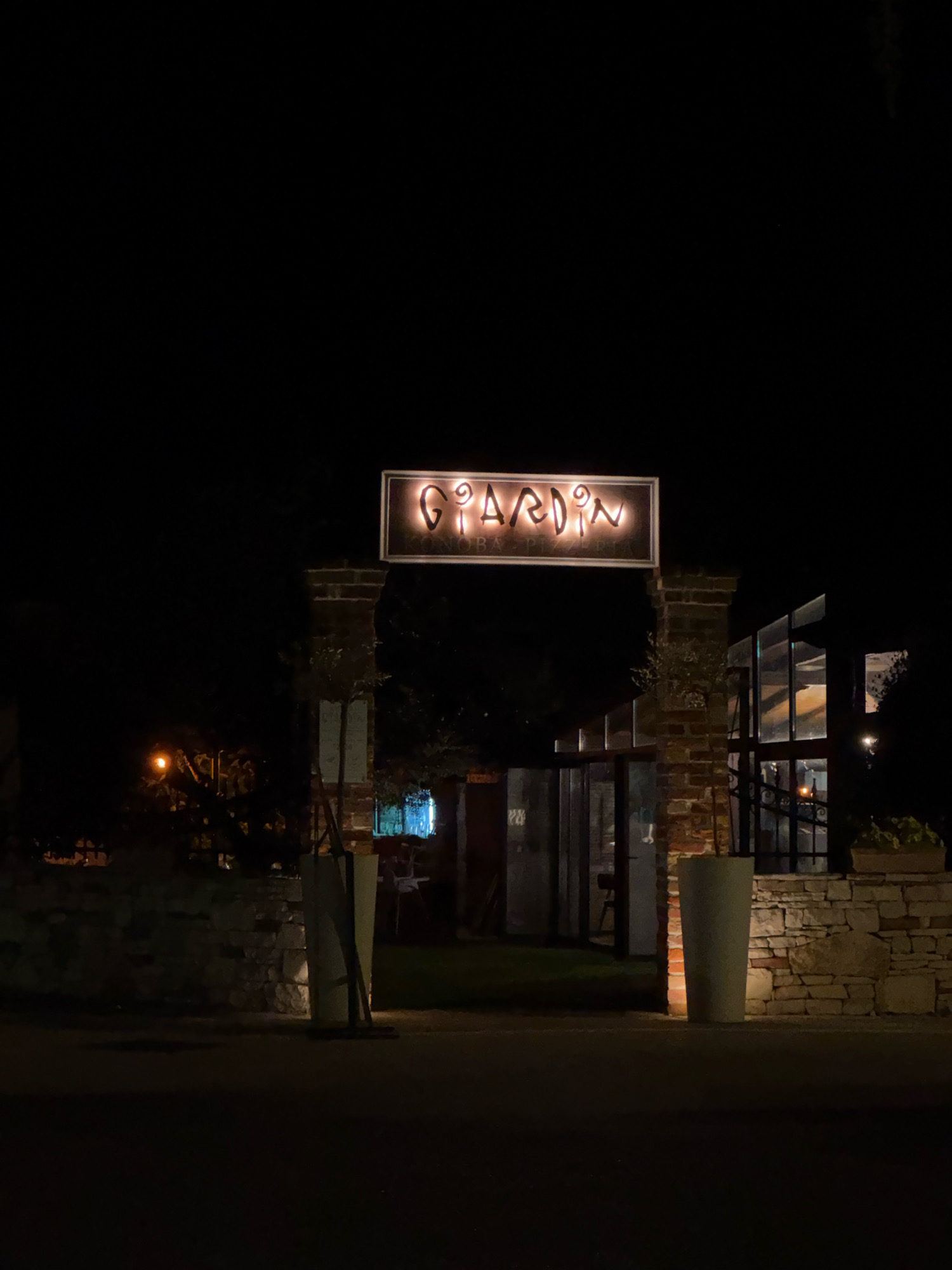 The image is a night-time exterior shot of a restaurant called "GIARDIN" located in the city of Novigrad, Croatia. The restaurant's sign, illuminated and featuring stylized lettering, is prominently displayed above an entranceway. The entrance is flanked by brick pillars and features large glass doors. To the right of the entrance, there is a modern building with large glass windows, revealing glimpses of the interior. The foreground shows a paved area, possibly a street or pathway, with large planters on either side of the entrance. A low stone wall runs along the left side of the image, with vegetation visible behind it. The overall atmosphere is dark, with artificial lighting highlighting the restaurant's signage and interior.