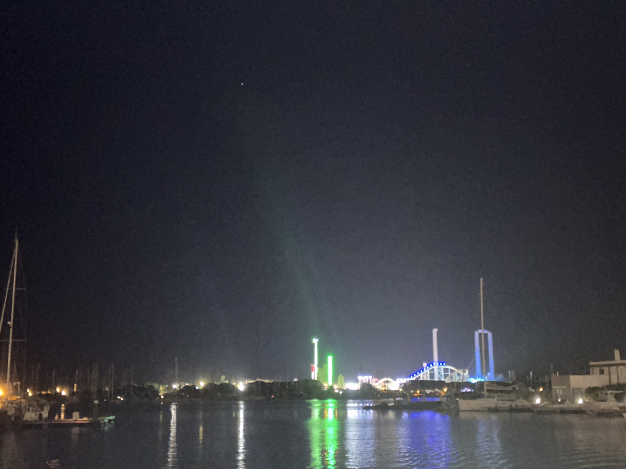 The image captures a nighttime view of a harbor in Agde, France, likely near the marina or a waterfront amusement area. The dark sky is punctuated by a single bright star or distant light in the upper left quadrant. The water in the foreground reflects the lights from the shore, creating shimmering streaks of color.
On the left side of the frame, several sailboats are docked, their masts reaching into the night. Their lights cast a warm glow on the water. In the middle ground, a collection of amusement park rides is illuminated by vibrant, colorful lights, predominantly green and blue. These rides appear to be tall structures, possibly roller coasters or observation towers, with bright vertical beams of light. A Ferris wheel or similar circular structure with blue lights is visible in the background.
To the right, a building with a white facade and a beige roof is partially visible, suggesting a residential or commercial area along the waterfront. The overall scene depicts a lively nighttime atmosphere with a blend of maritime activity and entertainment.