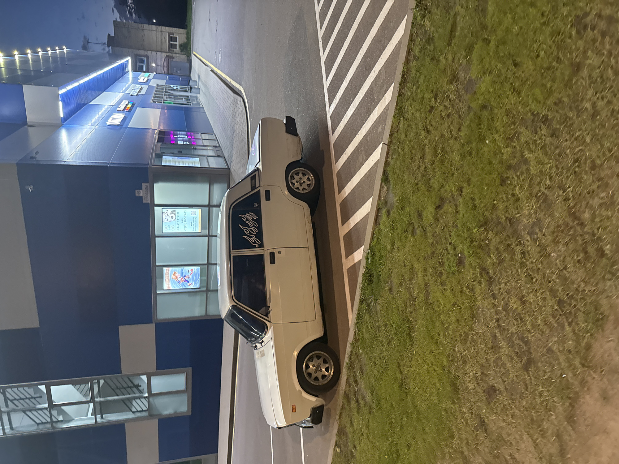 A white four-door sedan is parked next to a modern building with blue and white panels, and bright fluorescent lighting. The car is positioned on an asphalt surface with white diagonal lines painted on it, suggesting a parking area. The building appears to be a commercial establishment, with multiple large windows displaying advertisements and illuminated signage. One sign reads "ОПТИКА" (Optika), indicating an optical store. Adjacent to the building, there is a sidewalk and a patch of green grass. The sky is dark, indicating it is either dusk or night. The scene is devoid of any visible people or ongoing activity. The car has a decal on the rear side window that resembles flames. The location is identified as Krasnoye Selo, Russia.