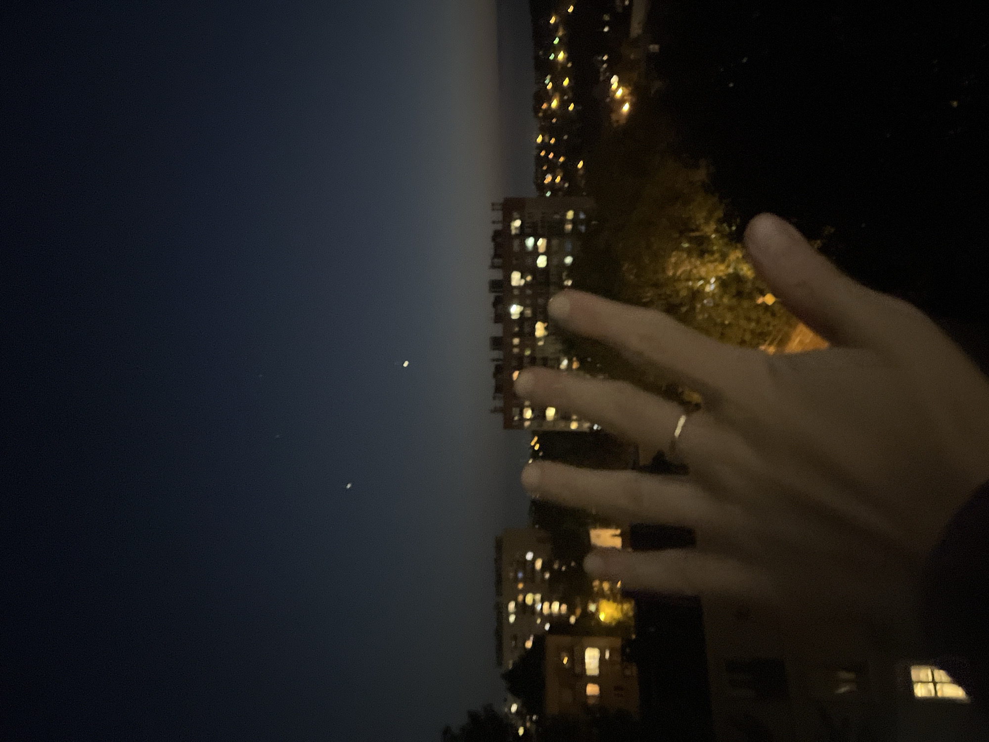 The image captures an urban nighttime scene. The left side displays a dark sky with two small, indistinct bright spots. Adjacent to the sky, a vertical arrangement of buildings with illuminated windows suggests a densely populated area. A human hand is visible in the foreground, positioned in front of the buildings and partially obscuring the view. The right side is darker with indistinguishable shapes of trees and other structures. It appears to be taken from within a building, since there is a visible window in the lower right. The location is unspecified in the city of Unknown, Unknown.