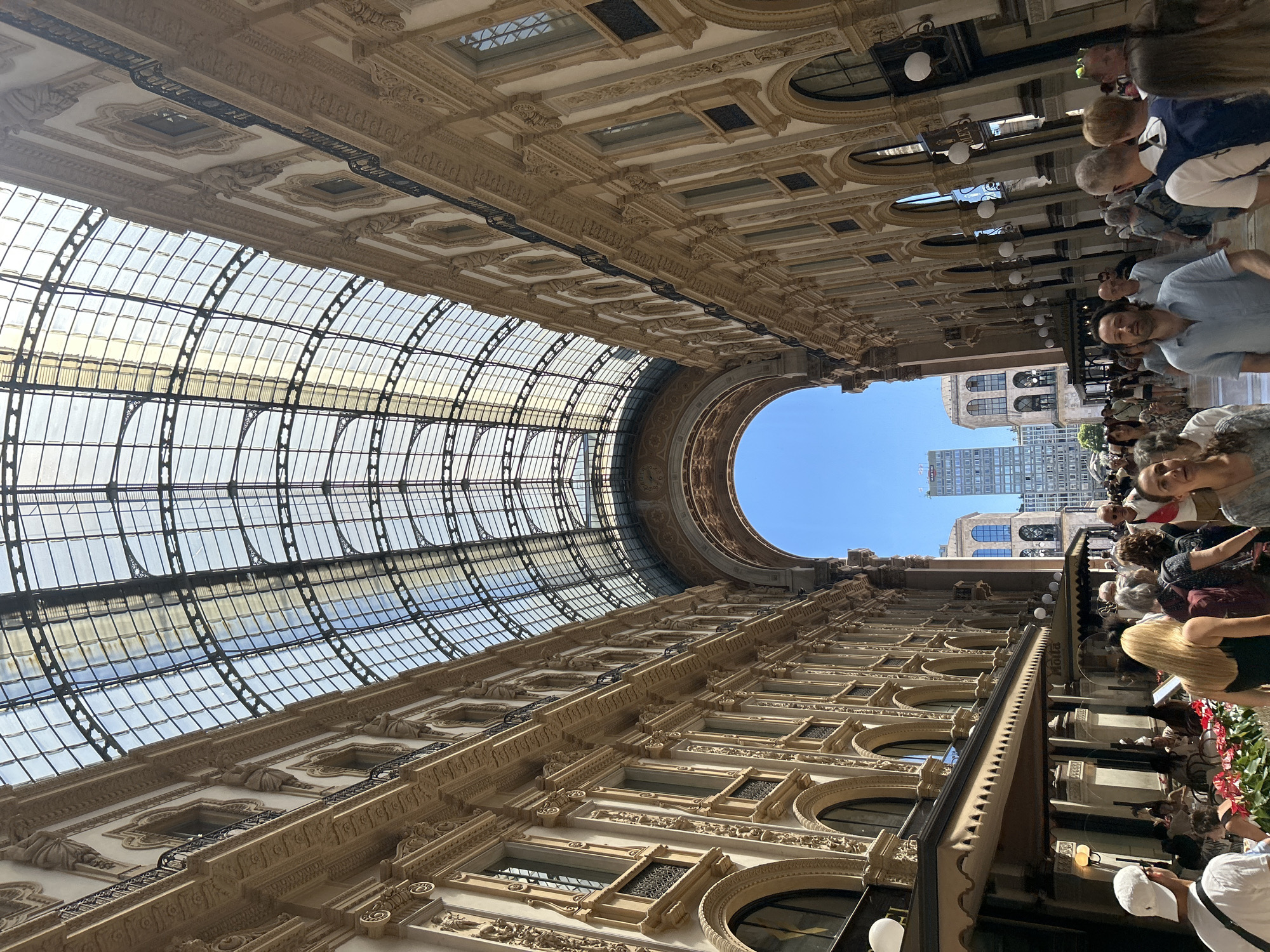 This media file presents a captivating upward view within a grand, ornate public space, strongly suggestive of the Galleria Vittorio Emanuele II in Milan, Italy. The dominant feature is the vast, arched glass and iron ceiling that forms the arcade's roof. Intricate dark metalwork supports numerous transparent glass panels, allowing abundant natural light to flood the interior.
At the far end, the glass ceiling culminates in a large, circular oculus that is open to the sky, revealing a clear blue expanse and the distant outlines of modern high-rise buildings, providing a juxtaposition of old and new urban architecture.
On either side of the arcade, multi-story buildings rise, showcasing elaborate classical architecture. Their facades are constructed from light-colored stone, richly decorated with ornate carvings, cornices, balustrades, arched windows, and decorative moldings, reflecting a sophisticated 19th-century design. Various light fixtures are also visible along the upper levels of these structures.
Numerous people populate the lower level of the arcade, appearing as a bustling crowd of tourists and shoppers. Dressed in casual daytime clothing, they are seen walking, standing, and observing their surroundings. In the foreground on the right, several individuals are partially visible, including a man in a light blue polo shirt and a woman with blonde hair. Hints of shopfronts, possibly adorned with red floral decorations, can be seen along the lower right side.
The bright light streaming through the glass ceiling and the clear sky visible through the oculus indicate it is daytime under fair weather conditions. The scene conveys a lively and relaxed atmosphere, with people enjoying the historical and architectural splendor of the gallery. No distinct text is clearly legible on any signs.