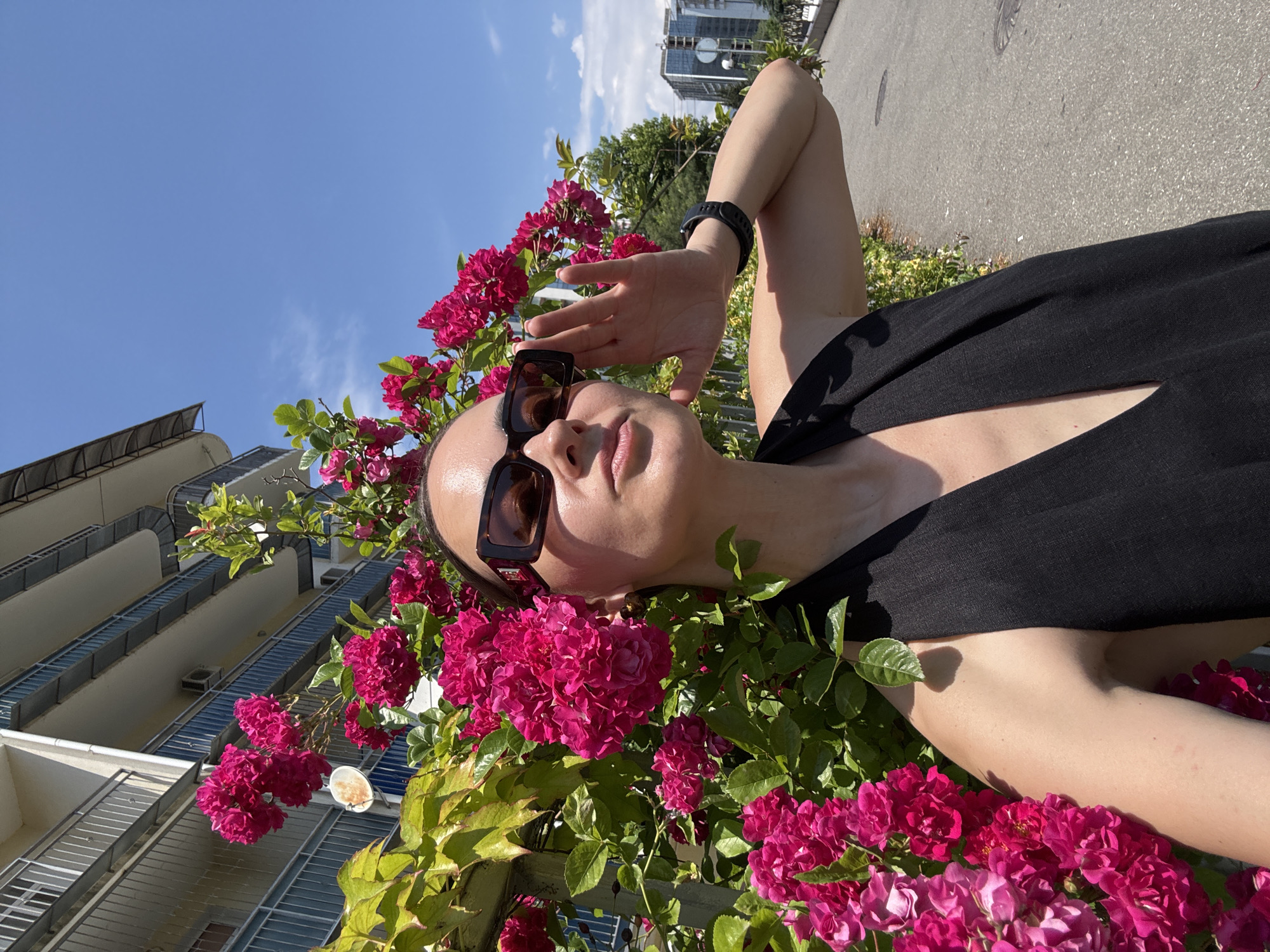 A woman in sunglasses and a black halter top stands beside a vibrant pink rose bush against a backdrop of a multi-story building with balconies and a bright blue sky. She is looking upwards, with her right hand raised to her face. The scene appears to be in an urban setting, with a paved walkway and road visible to the right, and modern buildings in the distance. The overall impression is a sunny day in a city.