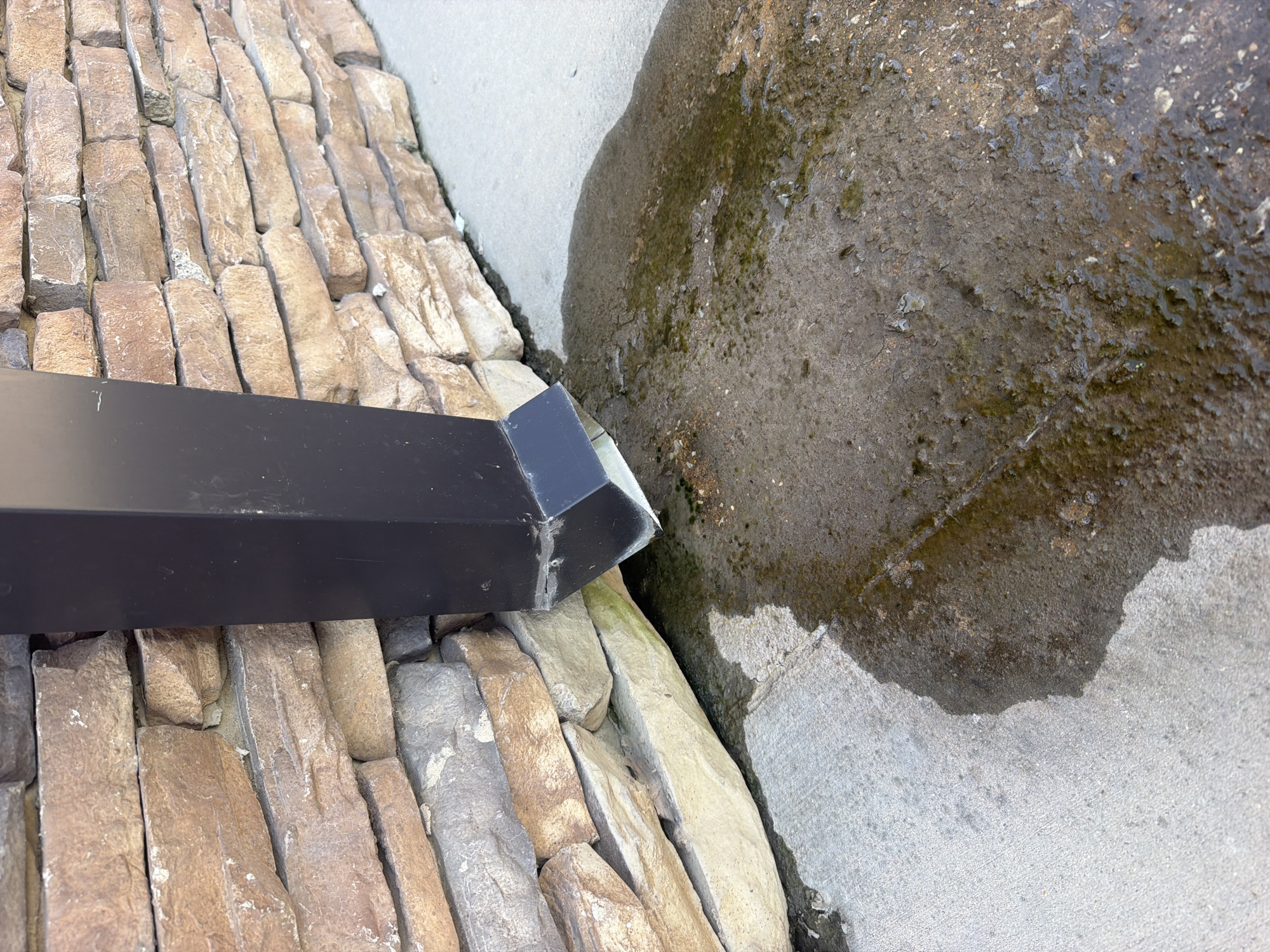 A black downspout extends from an unseen source above and directs water onto a textured concrete surface. The downspout is positioned against a wall constructed of stacked, rough-hewn, tan and brown stones. A puddle of water has formed on the concrete, extending outwards from the downspout's outlet and appearing somewhat murky with traces of green algae or moss. The scene appears to be outdoors, likely on the exterior of a building in Cardwell, United States. There are no people or animals visible in the image.