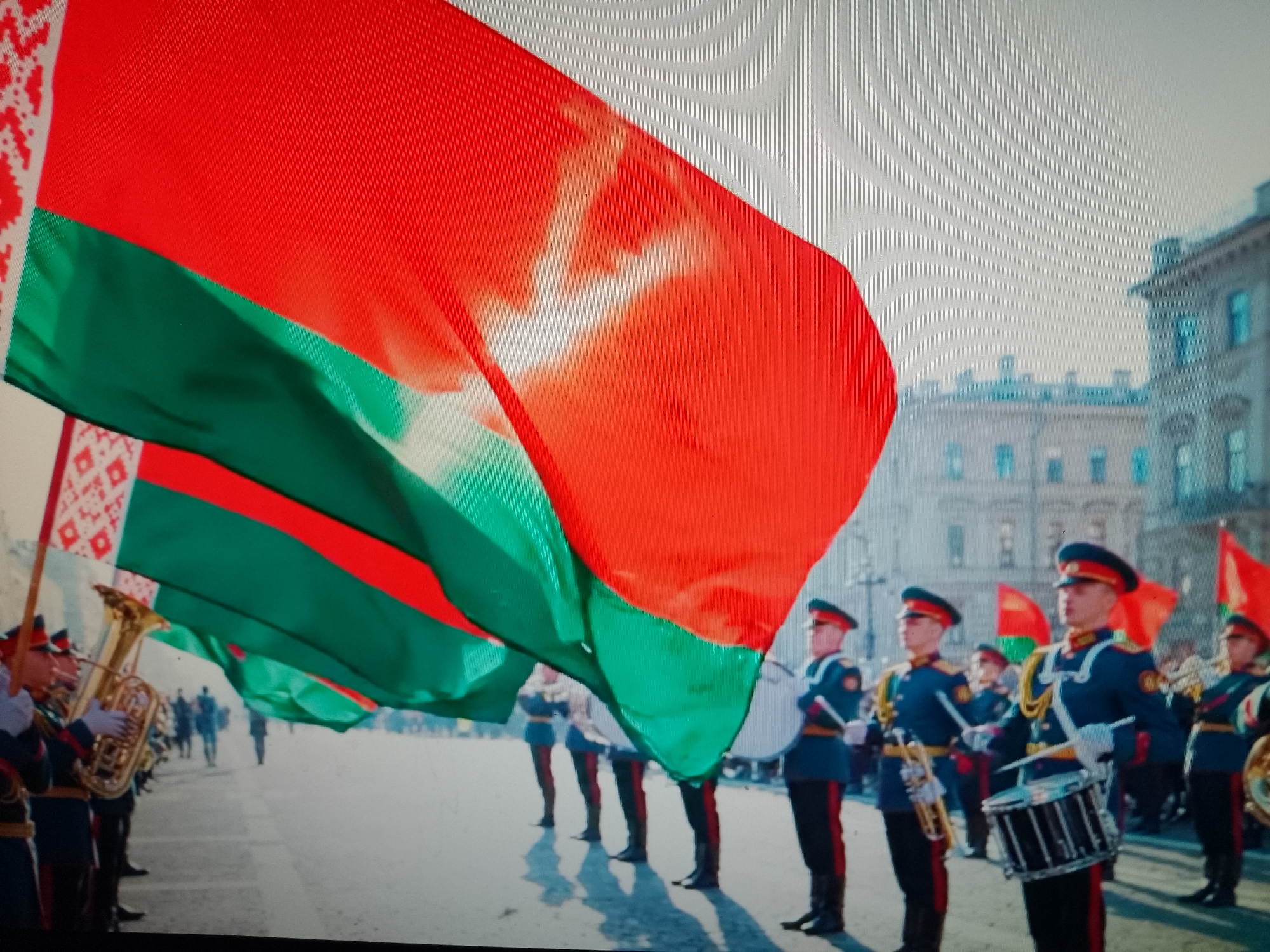 In an outdoor urban setting, a group of people in military uniform stand in formation. They are dressed in dark blue jackets with red trim and red hats. Some individuals hold musical instruments, including tubas and drums. Multiple large flags are being displayed, featuring red and green horizontal stripes with a white ornamental pattern on the left side. The flags appear to be unfurled and are the focal point of the image. In the background, there are buildings with architectural details and windows, suggesting an urban environment. The sky is visible at the top of the image. The ground appears to be paved. The presence of a military band and flags implies a ceremonial event or parade. The location is stated as Chiajna, Romania.