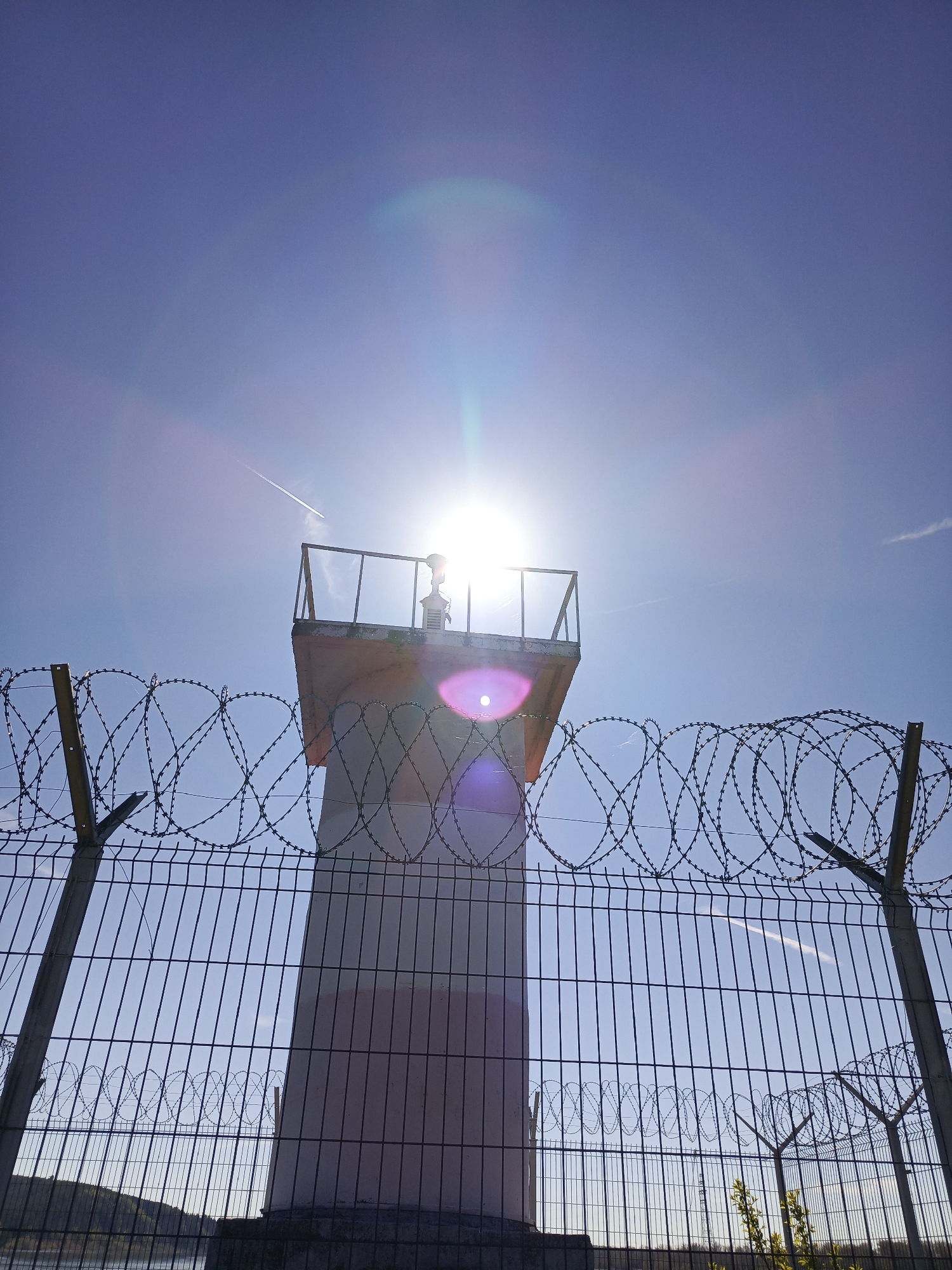The image shows a watchtower behind a chain-link fence topped with barbed wire. The tower is primarily white, with an orange-tinted top section and a small platform with railings. The sun is directly behind the tower, creating a lens flare effect. The sky is blue and mostly clear. The fence and barbed wire suggest a security perimeter. A trace of an airplane can be seen crossing the image in the upper right. The bottom left corner contains a patch of land and water. According to image information, this image was taken in Chaikovsky, Russia.