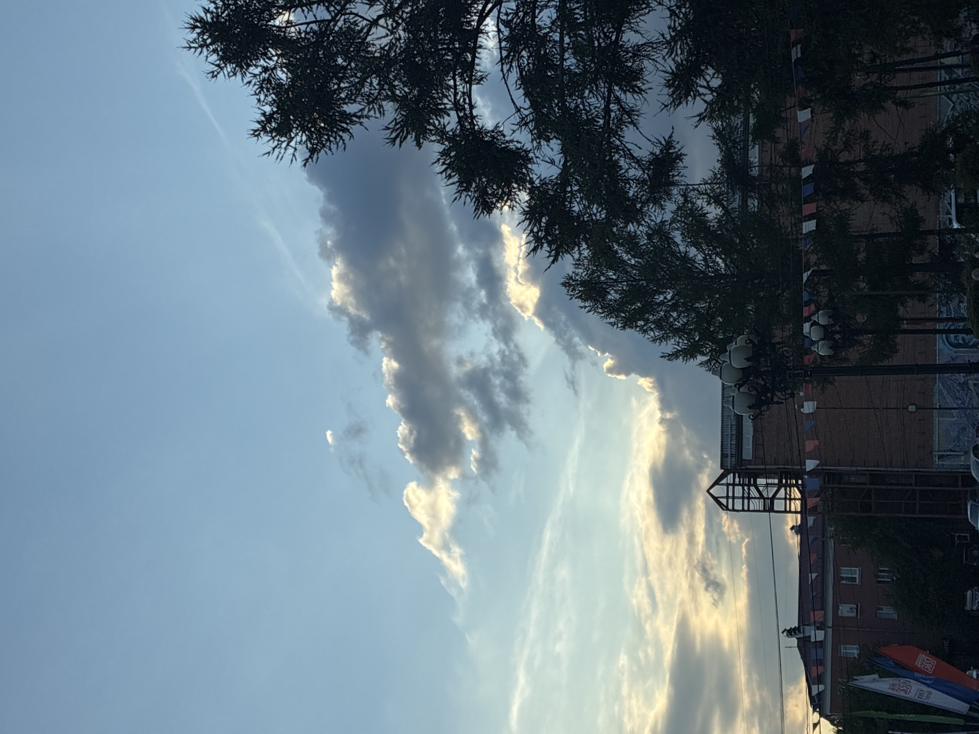 The image captures a view of the sky and buildings in Irkutsk, Russia. The sky is predominantly blue with scattered clouds, some of which are illuminated by the sun. The clouds display a gradient of light and shadow, with bright edges where sunlight passes through. Silhouetted against the sky on the right side are the branches of several coniferous trees, their dark foliage creating a stark contrast.
Below the trees, buildings are visible. One building on the left appears to be red-roofed with multiple windows, suggesting it's a residential or older commercial structure. Adjacent to it, a taller, darker brick building rises, featuring an exposed metal framework structure on its upper section, possibly an architectural addition or an unfinished construction. A string of small, colorful flags, predominantly white, red, and blue, is strung across the foreground between lampposts. These flags are likely decorative or commemorative. Several streetlights with multiple globes are positioned along the front of the buildings. In the lower left corner, several flags are also visible, including white, red, and blue ones, some with lettering and logos. Cars are parked on the street in the lower portion of the image, some partially obscured by the flags and trees. The overall scene suggests an urban environment, possibly a public space or street, during daytime, with the setting sun casting dramatic light on the clouds.