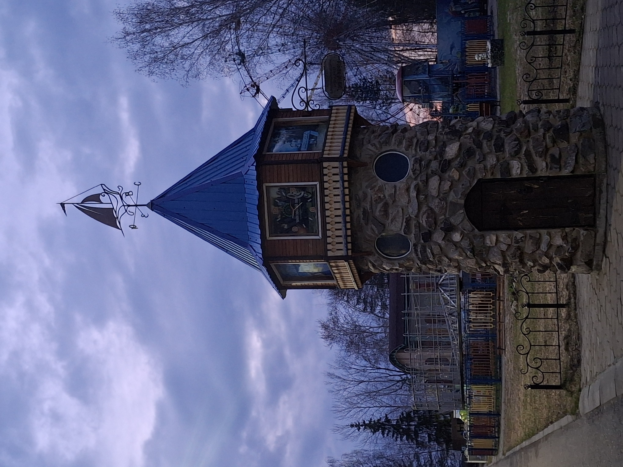 This media file captures a whimsical, fairytale-like tower situated within what appears to be an amusement or recreation park in Chaikovsky, Russia. The tower features a robust base made of irregular, rough-hewn stones, complete with a dark, arched wooden door and two small, round windows. Above the stone section, there is a wooden story with rectangular windows displaying colorful painted or printed scenes, including a clock face and other illustrative images. The tower is topped with a steep, blue conical roof that culminates in an ornate metallic weathervane shaped like a sailing ship.
The scene is set under an overcast sky, characterized by shades of gray and purple, suggesting either a heavily clouded day or the twilight hours of dusk or dawn. Bare, deciduous trees are visible in the background, implying late autumn, winter, or early spring. A paved walkway, constructed from dark pavers, runs alongside the tower. Beyond this path, a decorative black metal fence with intricate scrollwork elements marks the boundary of what appears to be a play area or a section of the park. In the deeper background, various structures associated with an amusement park, such as parts of rides and colorful playground equipment, are discernible. A small, dark sign hangs from a decorative iron bracket on the wooden section of the tower, but any text on it is not clearly legible. No people are present, and no specific activity is taking place, presenting a static view of the park's architectural features and environment.