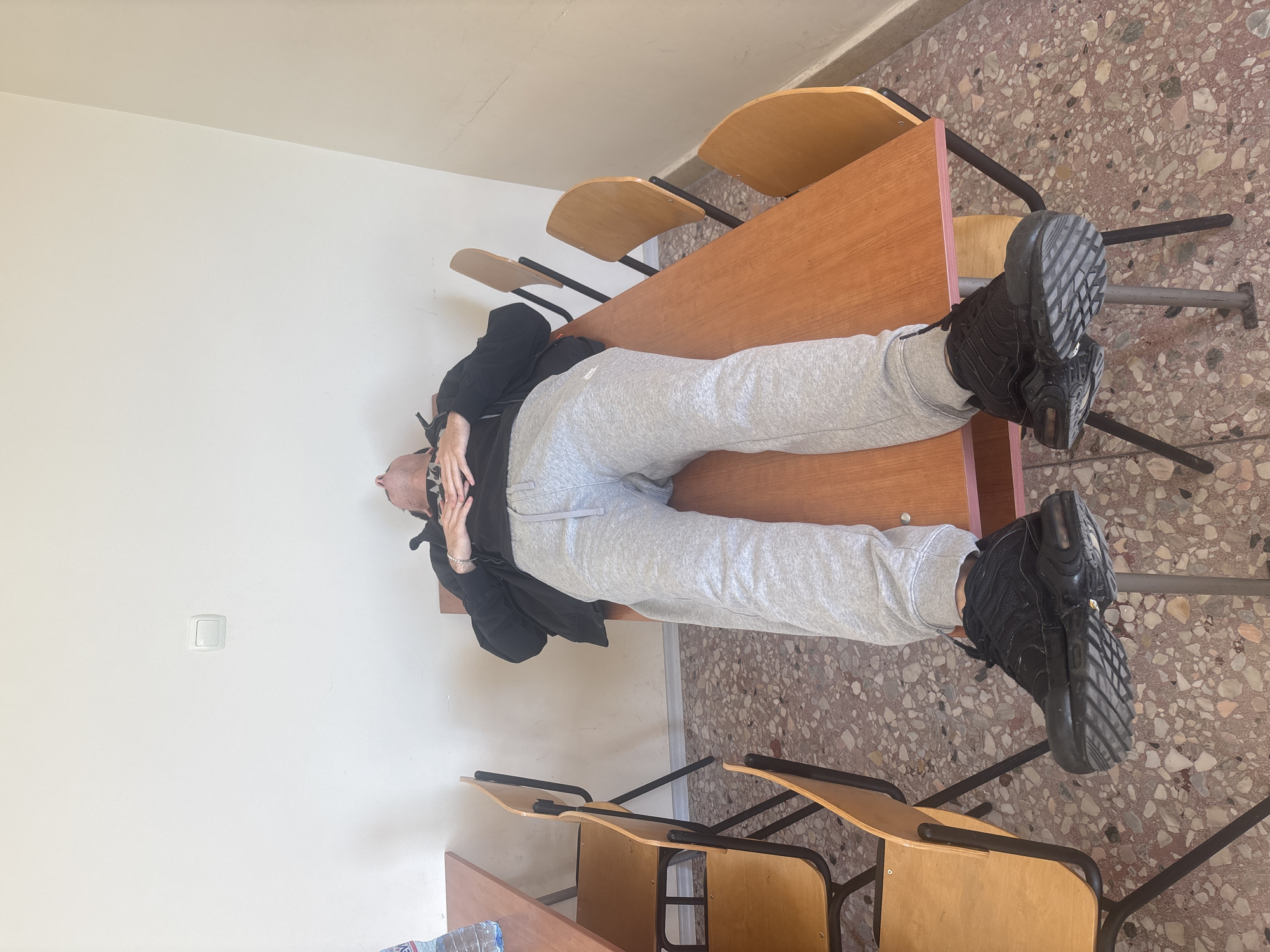 A person is lying face up on a wooden desk, legs extended over the edge and feet resting on chairs. The person is wearing gray sweatpants, black sneakers, and a dark jacket. The desk is part of a row of desks and chairs. The floor is covered with a speckled, terrazzo-like surface. A white wall is in the background, with a light switch visible.
The location appears to be a classroom in a school in Plovdiv, Bulgaria.