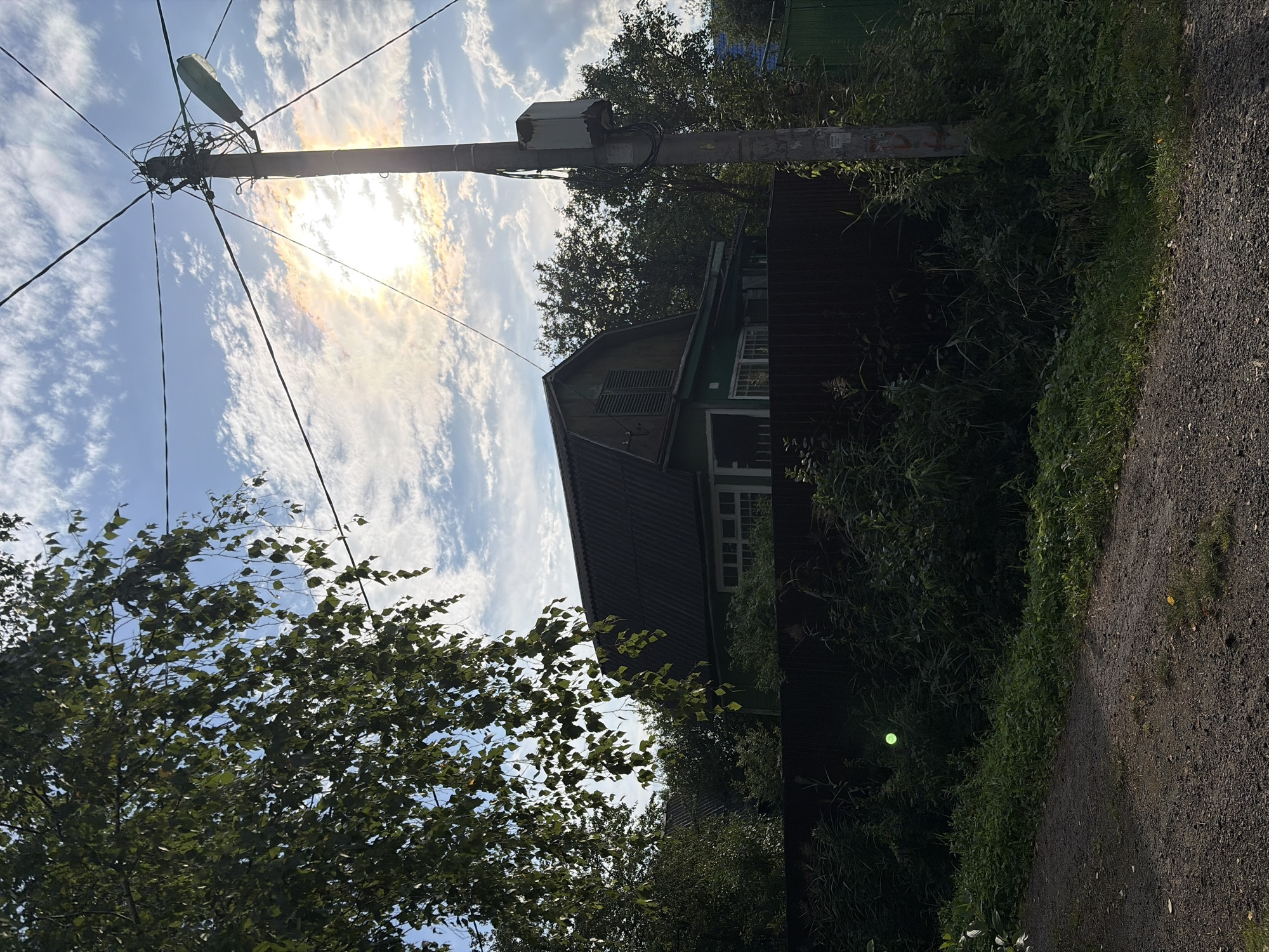 A sunlit view of a rural scene in Электротехник Снт, Russia, features a rustic green wooden house with a dark pitched roof, partially obscured by lush green foliage and trees. A dark wooden fence runs across the foreground. A concrete utility pole with overhead wires and a streetlight stands prominently to the right of the house. The sky above is a mix of blue with scattered white clouds, with the sun casting a bright glow through a section of clouds, creating a slight halo effect. A gravel path leads into the scene from the bottom left. A small, bright green, circular object is visible in the dense vegetation to the left of the house.
