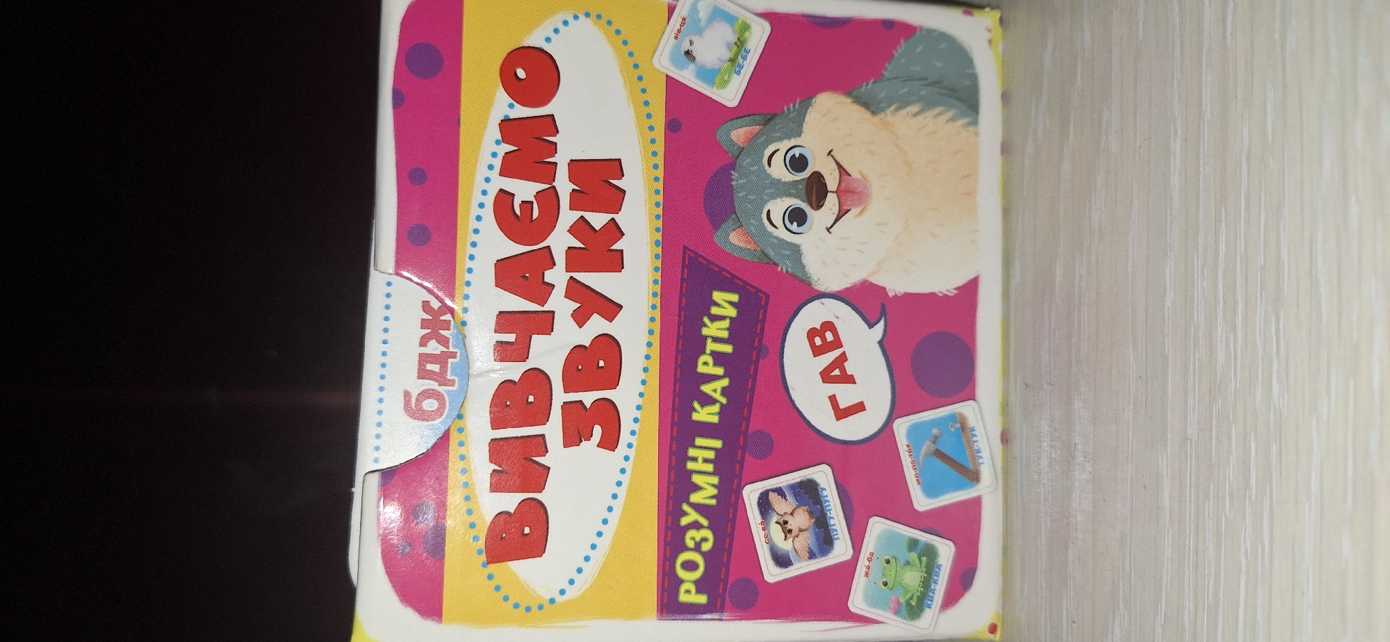 The image shows the front of a children's educational game box. The box features a cartoon wolf character and several small illustrated cards depicting animals and objects. The text on the box is in Ukrainian and appears to be related to learning sounds. The overall design is colorful and playful, typical for children's products. There is no identifiable information in the image that would place it in Bila Tserkva, Ukraine, or any specific city. The environment is a close-up shot of the box, likely on a table or flat surface, with a dark background behind it.
