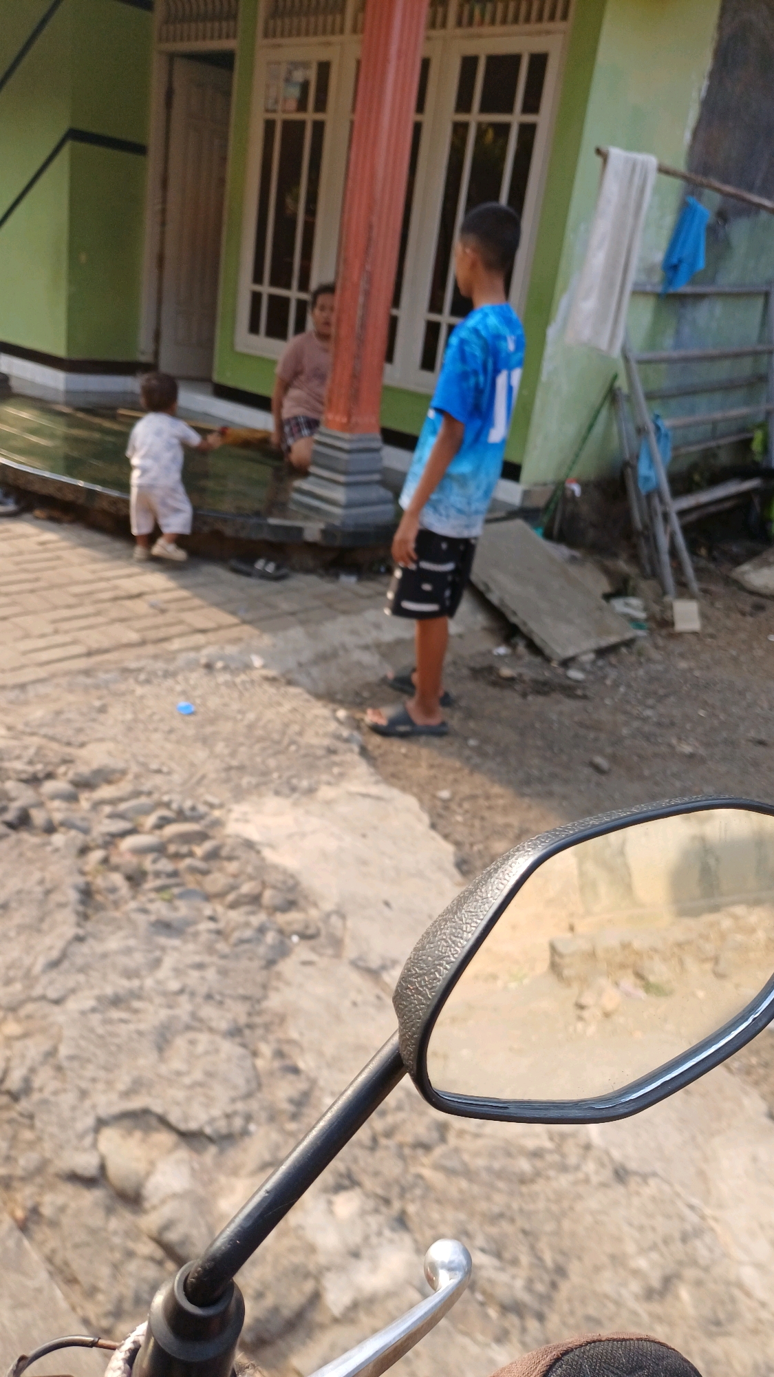 In Wonokerso, Indonesia, a scene unfolds on a paved and rocky outdoor area, likely a residential street, with a green and brown house visible in the background. A young boy, wearing a blue and white tie-dye t-shirt and black and white patterned shorts, stands facing away from the camera. Beside him, a younger child in a white outfit is walking away from the house towards the camera. In the background, an adult in a brown shirt and dark shorts is partially visible sitting near the house. The image appears to be taken from a motorcycle, with the handlebars and rearview mirror prominent in the foreground, suggesting a first-person perspective from a rider. The environment is sunny, with shadows cast on the ground.