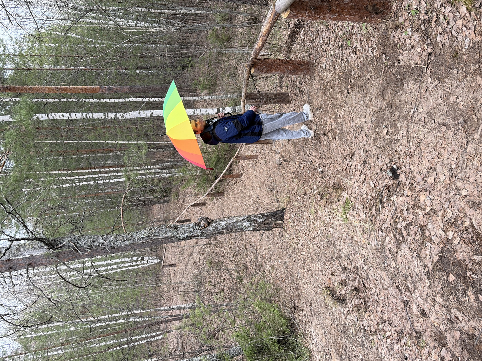 A man stands on a dirt path in a forest holding a multicolored umbrella. The path is covered in leaves and dirt. The trees are mostly bare, with some new growth visible. The man is wearing a blue jacket, light gray pants, and white sneakers. He is facing toward the forest with the umbrella open above his head. The location is the city of Chaikovsky, Russia.