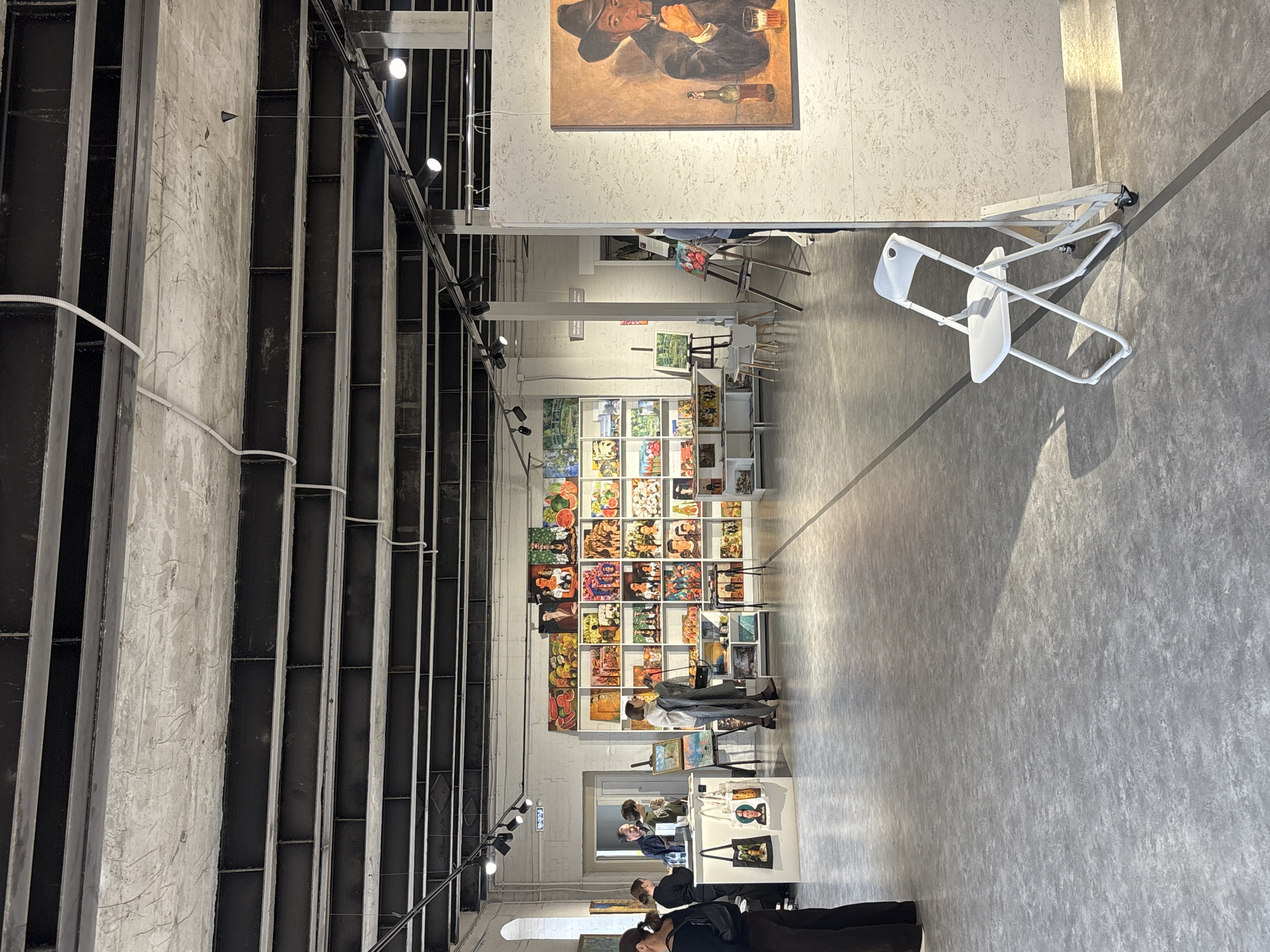 The image captures an interior scene in Krasnoyarsk, Russia, showcasing an art exhibition in what appears to be a converted industrial space. The room features a high ceiling with exposed metal beams and unfinished concrete walls, suggesting a contemporary aesthetic.
A large wall display is centrally located, filled with numerous framed artworks arranged in a grid pattern. Several visitors are present, some examining the art while others engage in conversations or browsing displays. Display tables showcase smaller items, possibly prints or related merchandise.
Notably, a painting of a man in a hat drinking beer hangs near the entrance. A white folding chair sits near the wall.