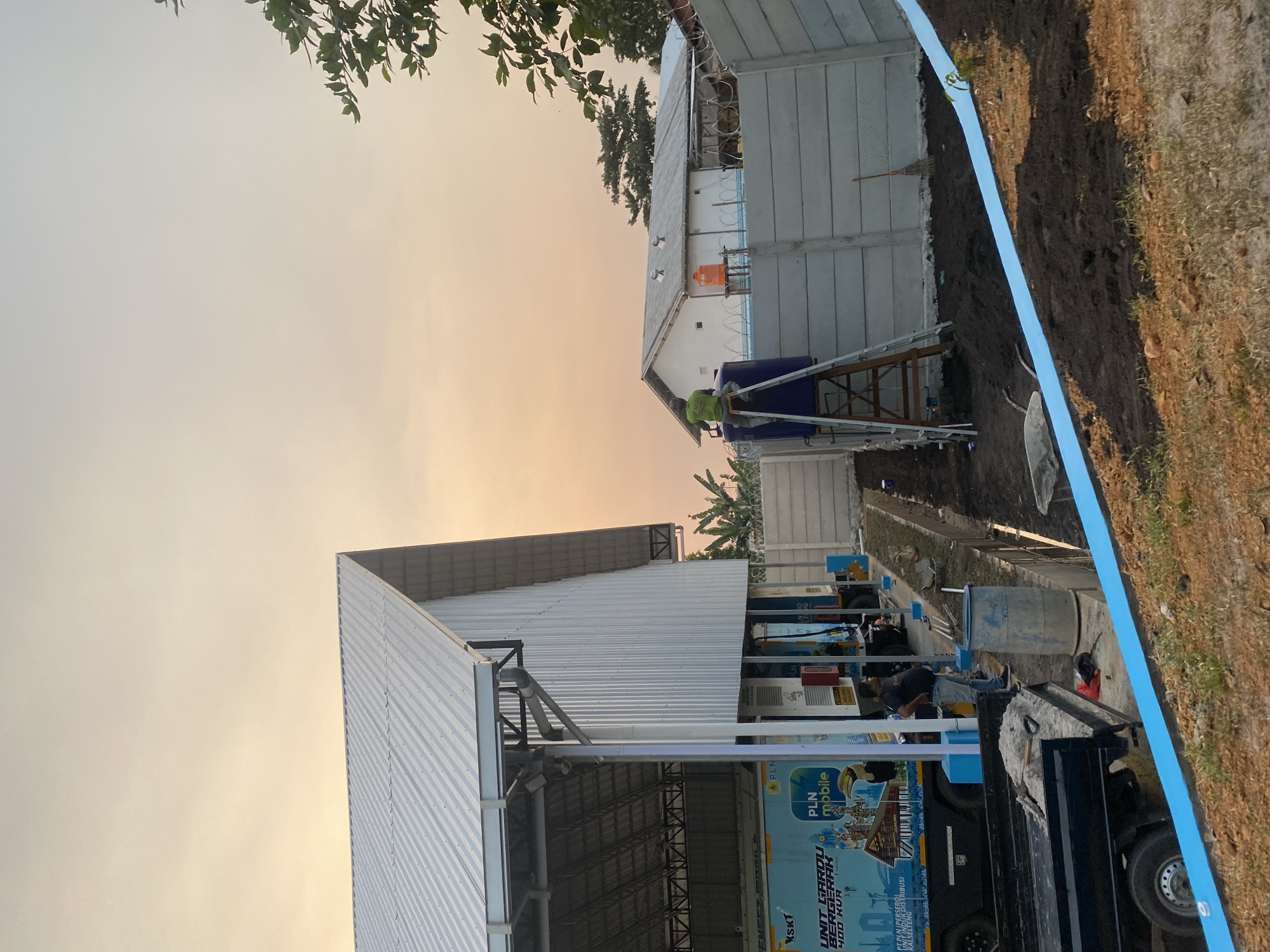 The image depicts an outdoor scene in the city of Banjarbaru, Indonesia, likely during sunset due to the warm, soft lighting and orange hue in the sky. In the foreground, there is a pile of dark soil and a blue barrier or tape running diagonally across the scene, suggesting ongoing construction or landscaping.
To the left, a modern building with a corrugated metal roof and white vertical siding is visible. Parked under this structure is a truck with a large blue graphic on its side, featuring text that includes "PLN mobile" and images of traditional Indonesian houses and a hornbill. Two individuals are standing near the truck.
To the right, a concrete wall with barbed wire on top separates the property from the street or another area. Beyond this wall, a smaller white building with a gray roof is situated. A person in a green shirt is standing on a ladder next to a large blue water tank, which is placed on top of another water tank. An orange water tank is also visible on a platform near the white building.
The overall impression is one of a construction site or industrial area undergoing some form of development or maintenance, with elements of the local Indonesian context evident in the truck's graphics and the tropical foliage visible in the background.