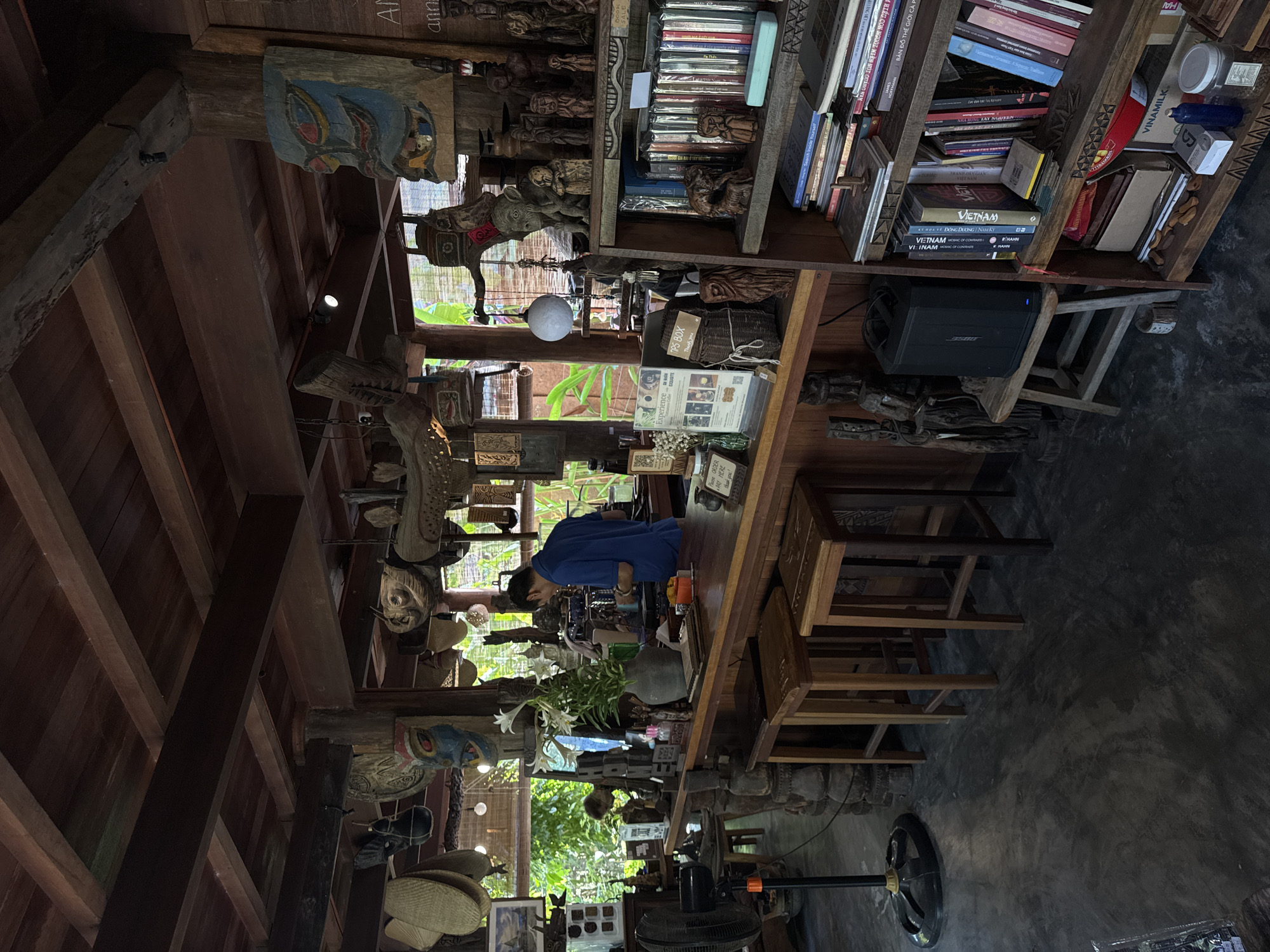 The image depicts the interior of what appears to be a shop or cafe in Đà Nẵng, Vietnam. The space is filled with an eclectic collection of items, including wooden carvings, books, and other decorative objects. A person wearing a blue shirt stands at a counter, possibly preparing drinks or serving customers. The interior features wooden beams and shelves, adding to the rustic aesthetic. Through the open windows, there's a glimpse of the outside greenery. A fan stands on the concrete floor. The shelves are stocked with books, some marked with "VIETNAM". Signage like "ORDER PAY HERE", "TIPS BOX Thank You", "Experience Exquisite" is visible.