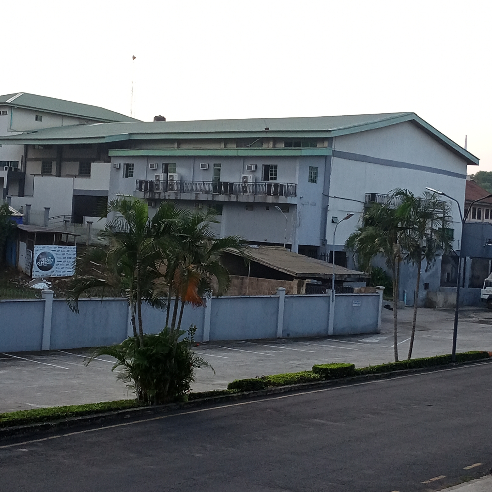 The image shows a building with a light blue exterior and a green roof, possibly an industrial or commercial complex, situated in Ibadan, Nigeria. In front of the building is a paved parking lot with white lines marking parking spaces. Palm trees and other greenery are visible in the foreground and around the property. A light blue wall with a decorative top borders the parking lot. A sign with "HOLY GHOST" and a circular logo is mounted on a smaller structure to the left, partially obscured by foliage. The sky above is overcast. The overall scene suggests an urban or semi-urban environment in Nigeria, likely during daylight hours.