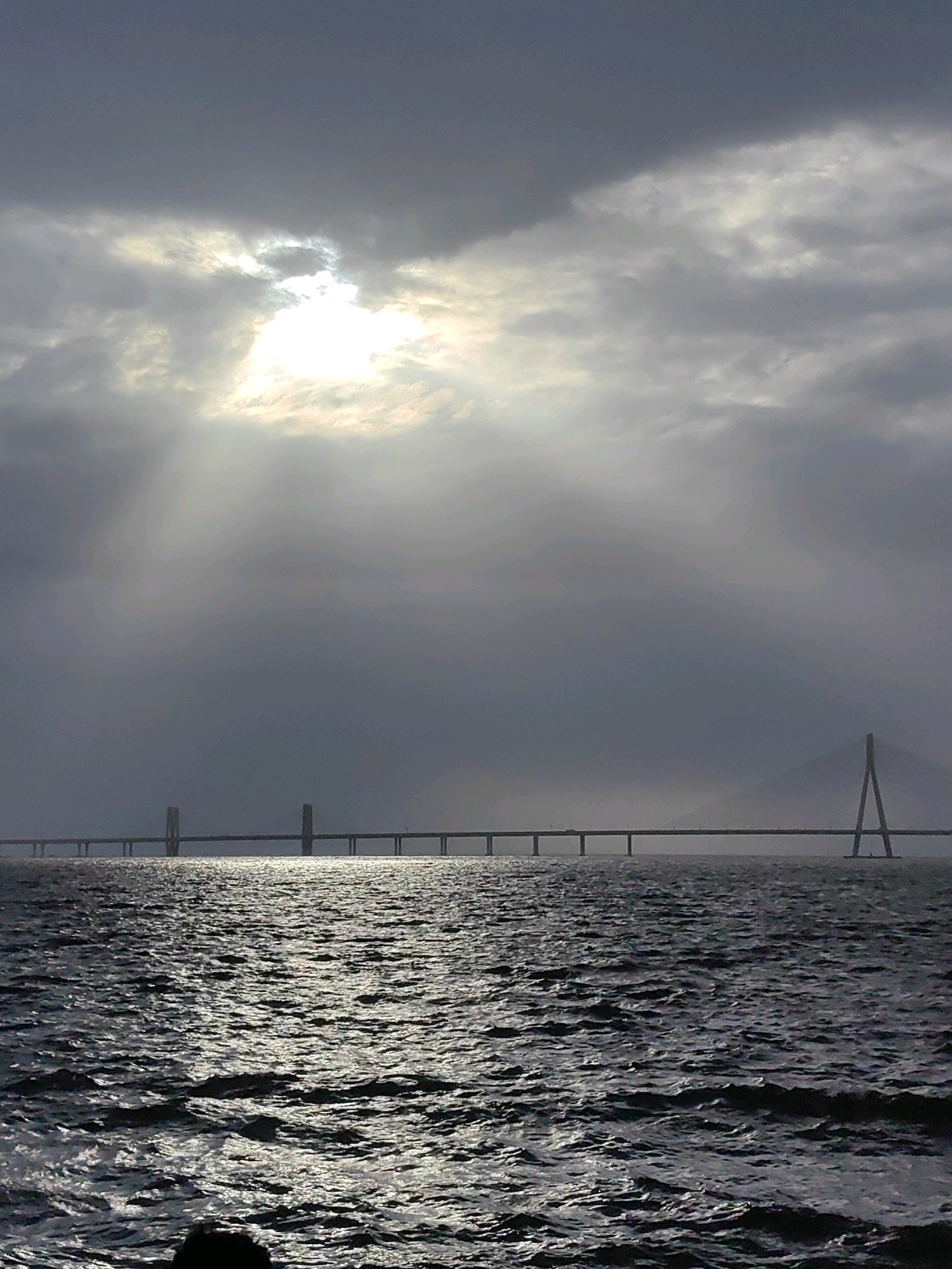 The image depicts the Bandra-Worli Sea Link bridge in Mumbai, India, under a cloudy sky with the sun peeking through the clouds. The sea is visible in the foreground, with choppy waves reflecting the light. The bridge stretches across the horizon, with its characteristic pylons and deck clearly visible. The overall atmosphere is dramatic and somewhat moody, with the interplay of light and shadow creating a sense of depth and scale. There are no people or vehicles immediately discernible on the bridge or in the water. The environment suggests a coastal setting within the city of Mumbai.