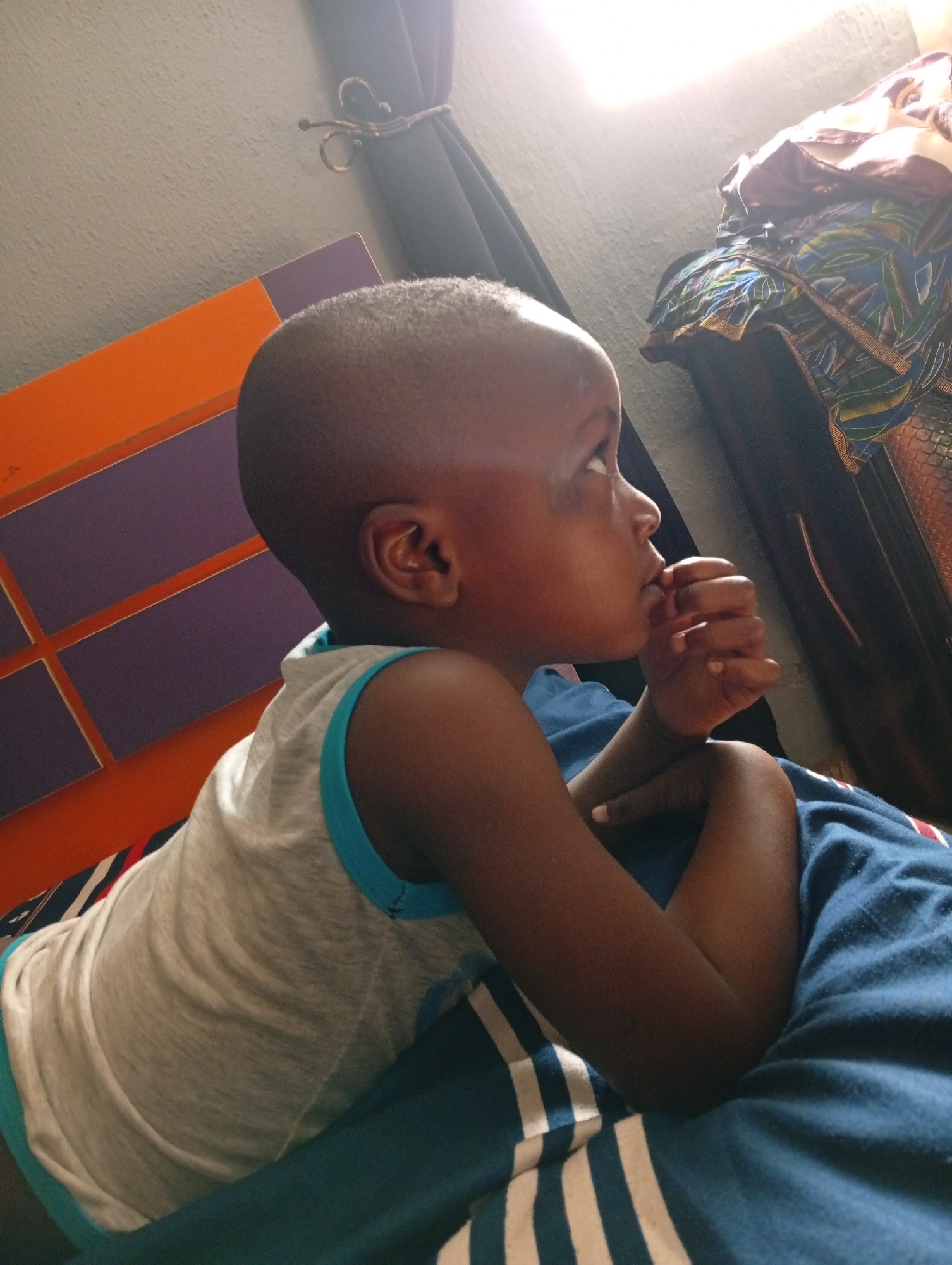 A young, dark-skinned child lies on a blue and white striped bed in a room. The child is wearing a gray tank top with blue trim. Their right arm is bent, and their hand is near their mouth. In the background, there is a headboard with orange and purple blocks. A dark curtain and part of an intricately patterned object stand beside the headboard. The light is bright in the scene, likely coming from a window. There is no way to determine the location, Sabon Tasha, Nigeria.