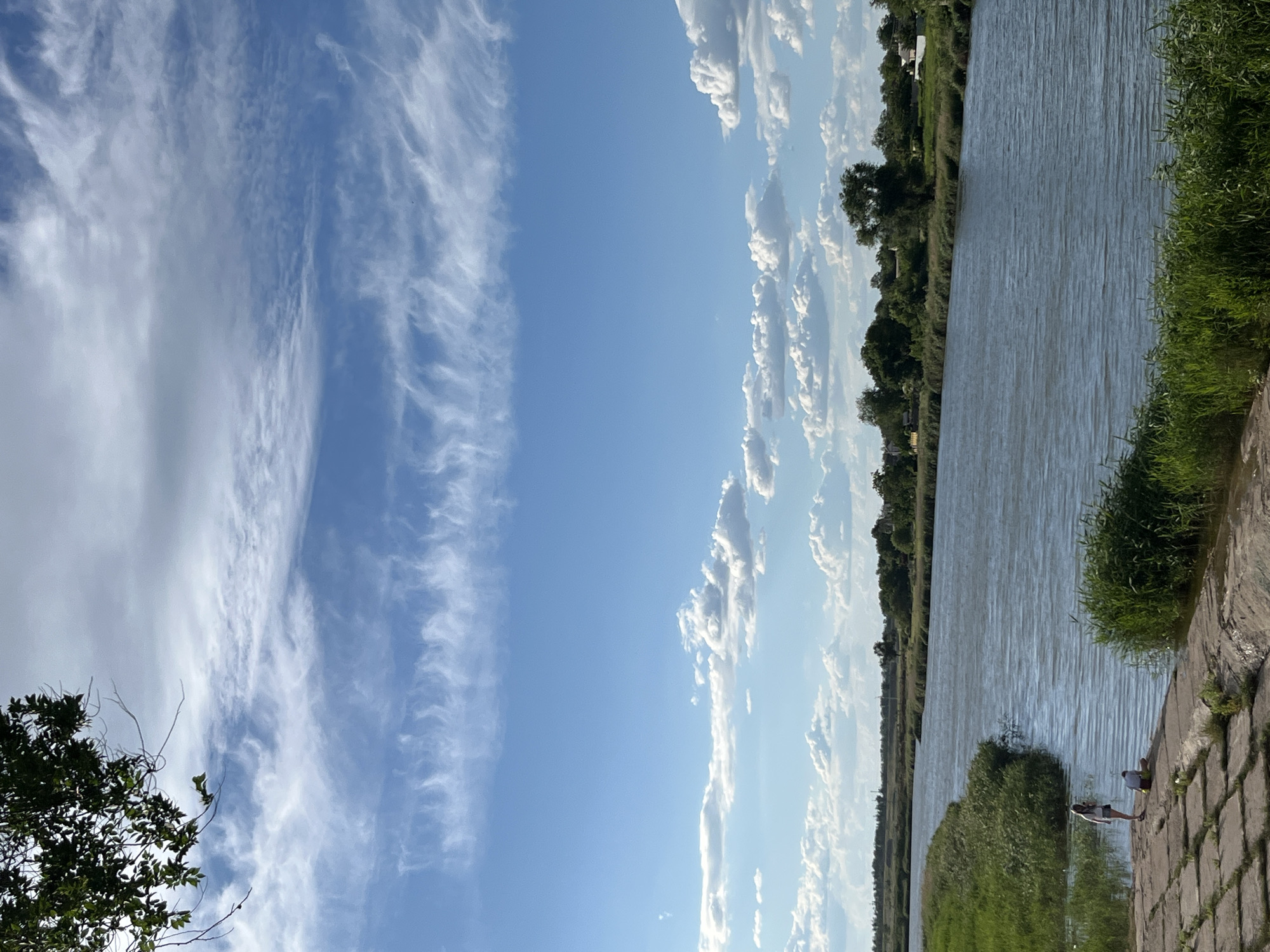 The image depicts a serene outdoor scene, likely during the daytime, given the bright sky and clear visibility. A wide river flows along the right side of the frame, its surface rippled by a gentle breeze. The opposite bank is lined with lush green trees and vegetation, suggesting a natural or park-like setting. The sky is a vibrant blue, dotted with various types of clouds, from wispy cirrus clouds stretching across the left side of the frame to more defined cumulus clouds lining the horizon.
On the paved embankment by the river, two individuals are visible. One person is seated, while the other stands nearby, both appearing to be enjoying the tranquil atmosphere. The embankment is constructed of large, rectangular stone slabs, adding a structured element to the natural landscape. The presence of the river, trees, and sky suggests a peaceful environment, possibly a recreational area or a scenic spot along a waterway. The overall mood is calm and relaxed, enhanced by the soft light and the gentle movement of the water and clouds. There is no visible text in the image.