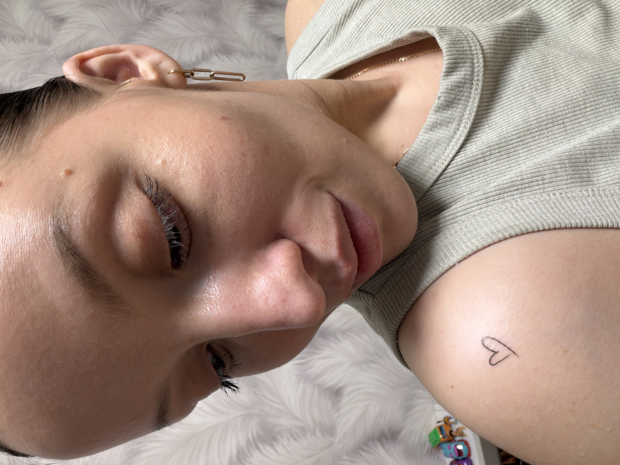 A close-up, high-angle shot shows a person lying down on a fluffy, light-colored surface, possibly a rug or blanket. The subject appears to be a young adult, with their eyes closed or partially closed, revealing long, dark eyelashes. Their skin is smooth with a mole visible near the ear and a small bump on the cheek. Dark hair is pulled back from their face.
The person wears a gold-colored, chain-link earring in their visible earlobe and a thin gold-colored chain necklace around their neck. They are dressed in a light green or sage-colored ribbed top or t-shirt. A small, outline heart tattoo is visible on their upper right arm or shoulder. Their lips are slightly parted, and their facial expression suggests a state of rest or relaxation. In the bottom right corner, a few small, colorful, toy-like objects are partially visible. The scene is brightly lit, indicating an indoor setting, likely a home in Reshetnikovo, Russia. There is no visible text in the image.