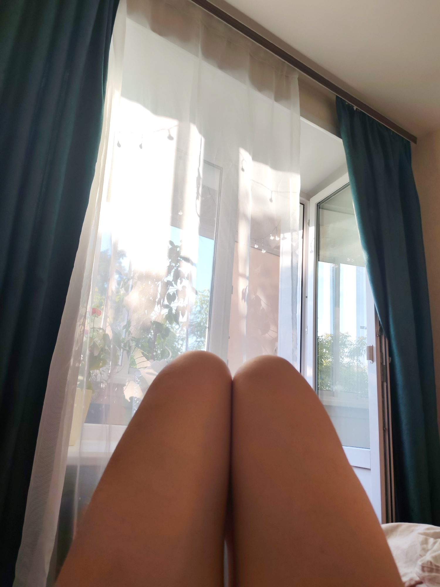 The image shows a person's legs, which are the primary subject, positioned in the foreground and taking up a significant portion of the frame. The legs appear to be bare and are slightly angled towards each other. Behind the legs is a window with sheer white curtains, allowing natural light to filter through. Through the curtains, glimpses of greenery and what appear to be potted plants are visible, suggesting an indoor setting with a view of the outdoors. To the left and right of the window are dark teal curtains, drawn partially closed. The overall lighting suggests it is daytime, with bright sunlight streaming in, creating a warm and slightly hazy atmosphere. The scene appears to be a bedroom or living room, indicated by the presence of curtains and a glimpse of bedding on the right side. The focus on the legs and the soft lighting evoke a sense of relaxation or a quiet moment. There is no visible text in the image. The location context is Izhevsk, Russia.